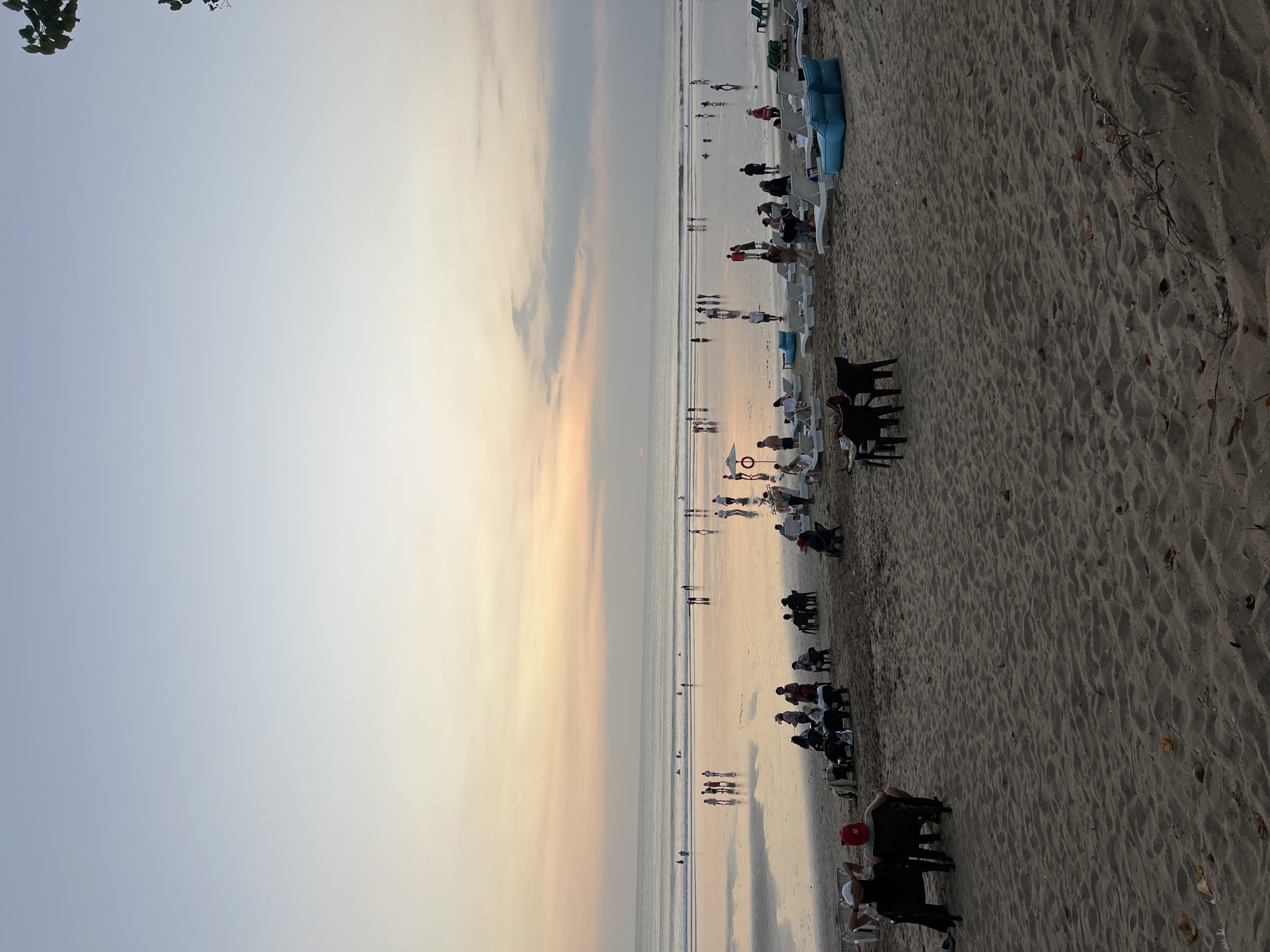 This image depicts a beach scene in Kuta, Indonesia, during what appears to be sunset or twilight. The sky is a gradient of pale blue at the top, transitioning to soft orange and pink hues near the horizon, with some scattered clouds. The ocean is calm, with gentle waves reaching the shore, and its surface reflects the colors of the sky, creating a shimmering effect.
On the sandy beach, numerous people are visible. In the foreground, several individuals are seated on chairs, some facing the ocean and others engaged in conversation. Further back, more people are scattered across the beach, some walking, others sitting on loungers or in groups. A collection of beach chairs and blue beanbag-style seating is arranged on the sand. In the distance, near the water's edge, a larger group of people are strolling or standing. The scene suggests a relaxed atmosphere, with beachgoers enjoying the evening ambiance. The sand in the foreground is textured with ripples and a few scattered leaves and debris. The overall lighting is soft and diffused, characteristic of the time of day.