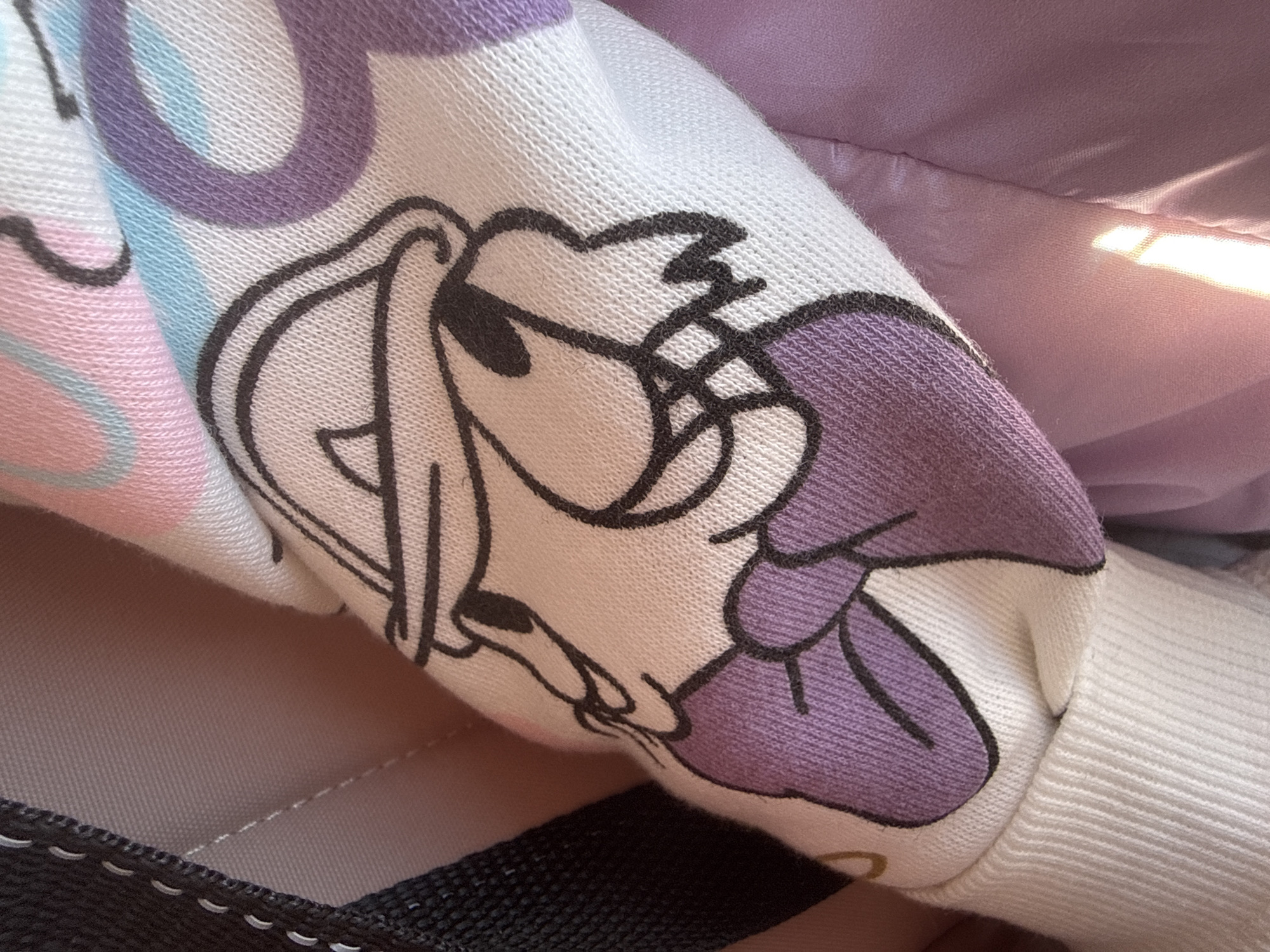 A close-up image shows a white sleeve with a print of Daisy Duck, rendered in purple and black outlines. The sleeve appears to be part of a sweatshirt or similar garment. The fabric has a textured, knitted appearance. In the background, partially visible, are a purple or lavender-colored material and what looks like a part of a black bag strap or handle. The setting is indeterminate and there is no information available in the image that indicates its location as Chaikovsky, Russia or any other specific place.