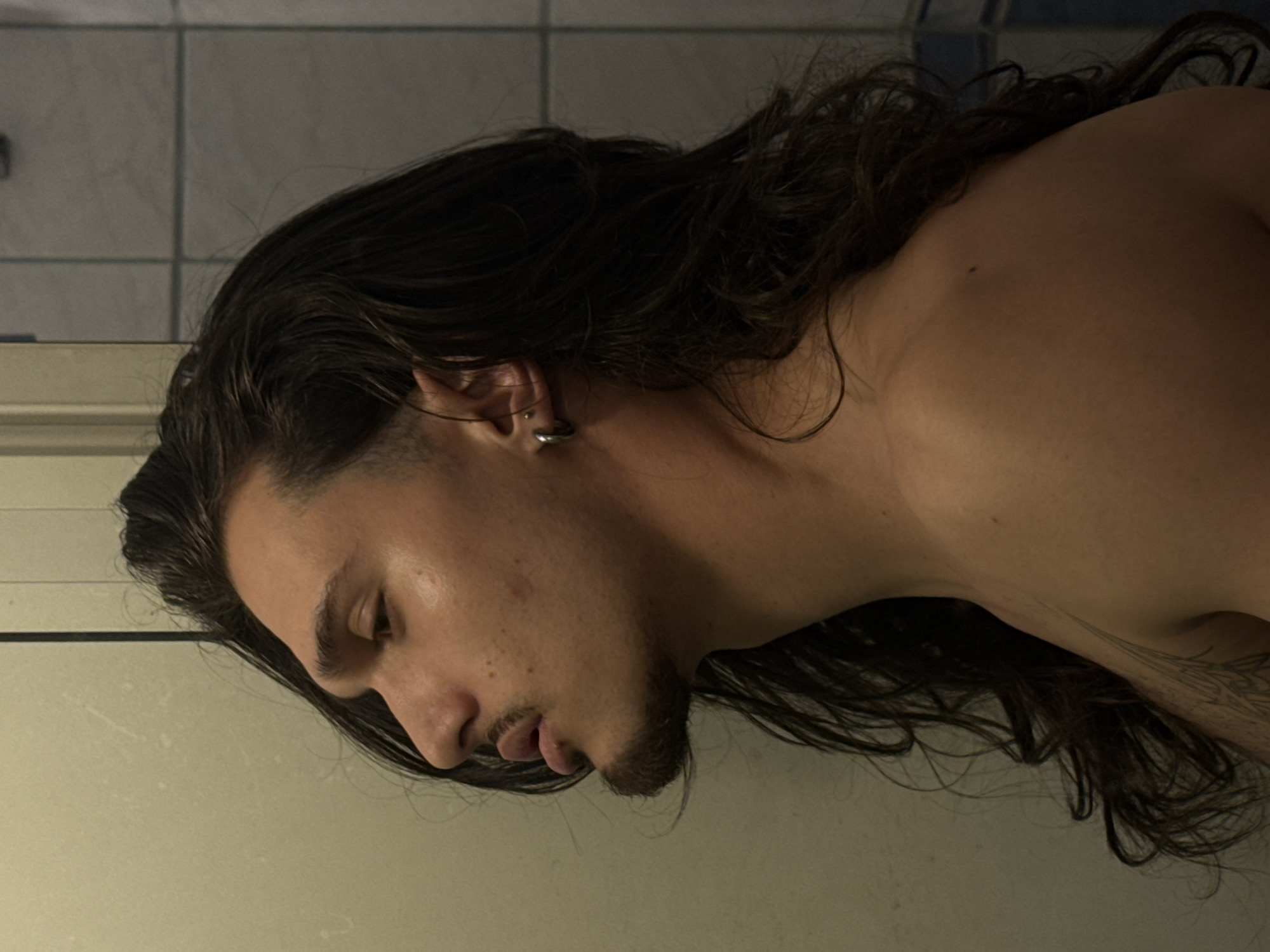 A young person with long, dark, wavy hair and a goatee is pictured in a profile view from the neck up. They have a small hoop earring in their left ear. The person's left shoulder is visible, and there is a tattoo on their left arm. The background consists of a light-colored wall and a tiled surface. The lighting is warm and appears to be artificial. There is no specific information within the image to indicate the location in Sofia, Bulgaria.