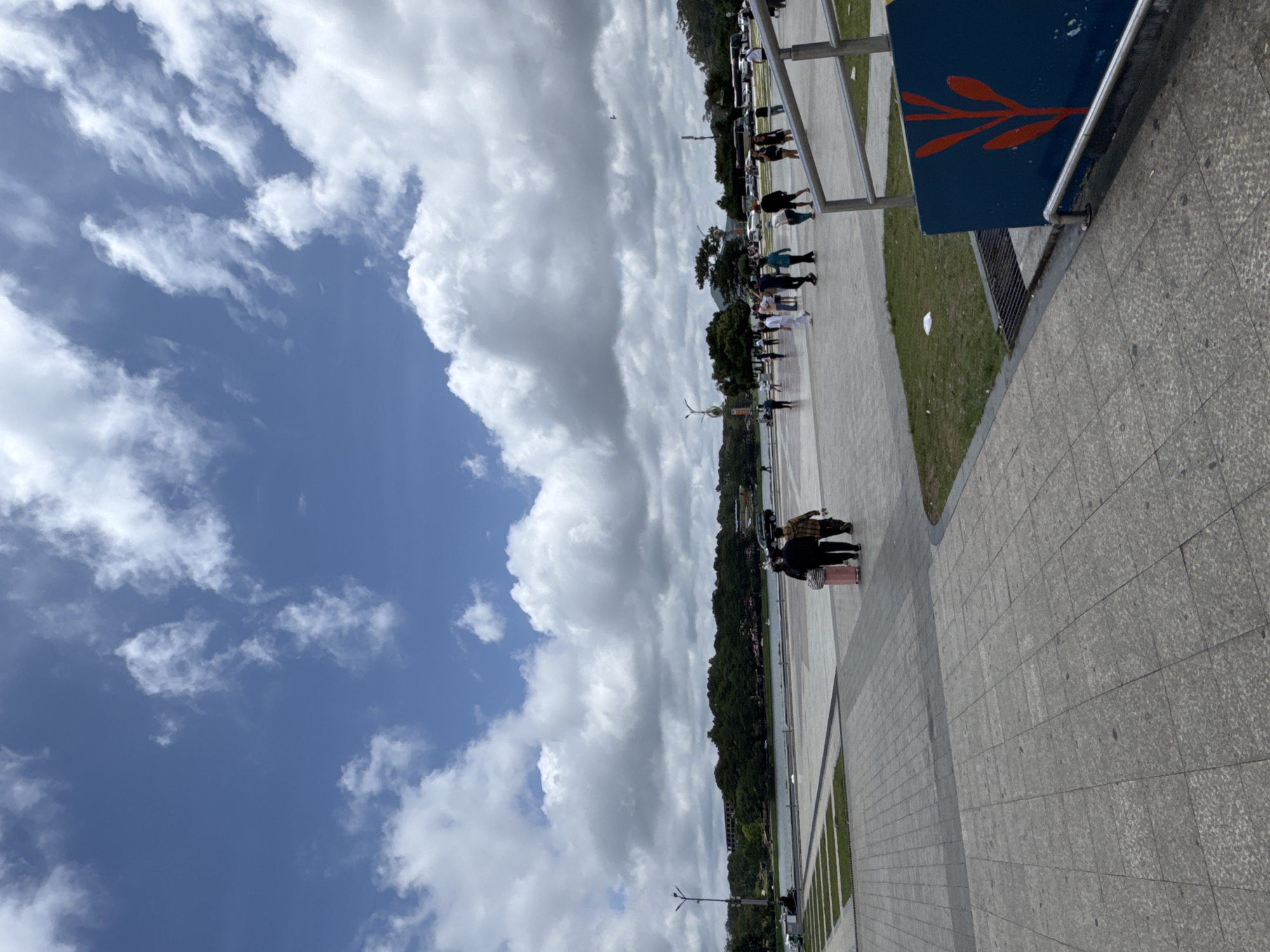 The image depicts a wide, paved public plaza in Da Lat, Vietnam, with a lake and lush greenery in the background. The sky is a vibrant blue, dotted with fluffy white clouds, suggesting a pleasant day. In the foreground, a couple is walking away from the camera, one pulling a pink suitcase. Further into the plaza, several other individuals and groups of people are seen strolling, engaged in casual activities. A dark-colored SUV and a bus are visible in the middle ground, hinting at the presence of transportation. The plaza is bordered by a railing and a blue structure with a red plant design, suggesting it's a well-maintained public space, likely near tourist attractions or a recreational area in Da Lat.
