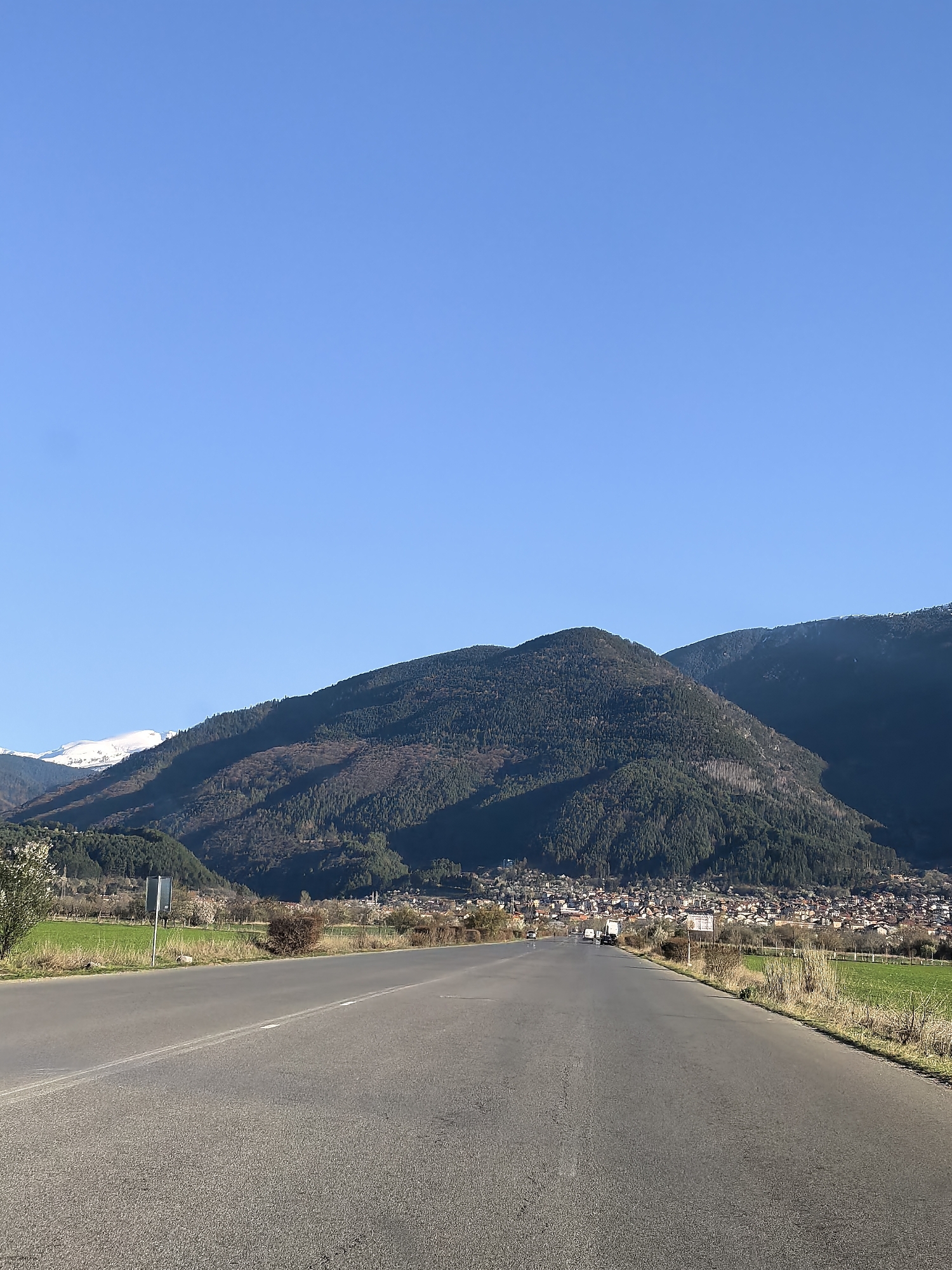 The image depicts a straight asphalt road leading towards a town nestled in front of a large, forested mountain range under a clear, blue sky. The road is flanked by fields and sparse vegetation on either side. There are a few vehicles visible on the road in the distance, approaching the town. The exact location within the city of Unknown, Unknown, is not discernible from the image.