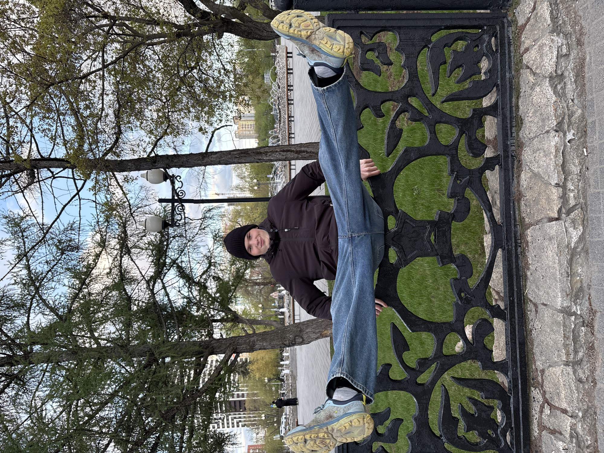 A person wearing a black hat and jacket and blue jeans is sitting on a black metal bench in Perm, Russia. The person's legs are extended upwards, with their shoes visible against the backdrop of trees and city buildings. The bench has a decorative design, and the surrounding environment includes trees, grass, and a stone pathway. In the background, fountains are visible. The photograph is taken at an unusual angle, with the image rotated 90 degrees counter-clockwise.