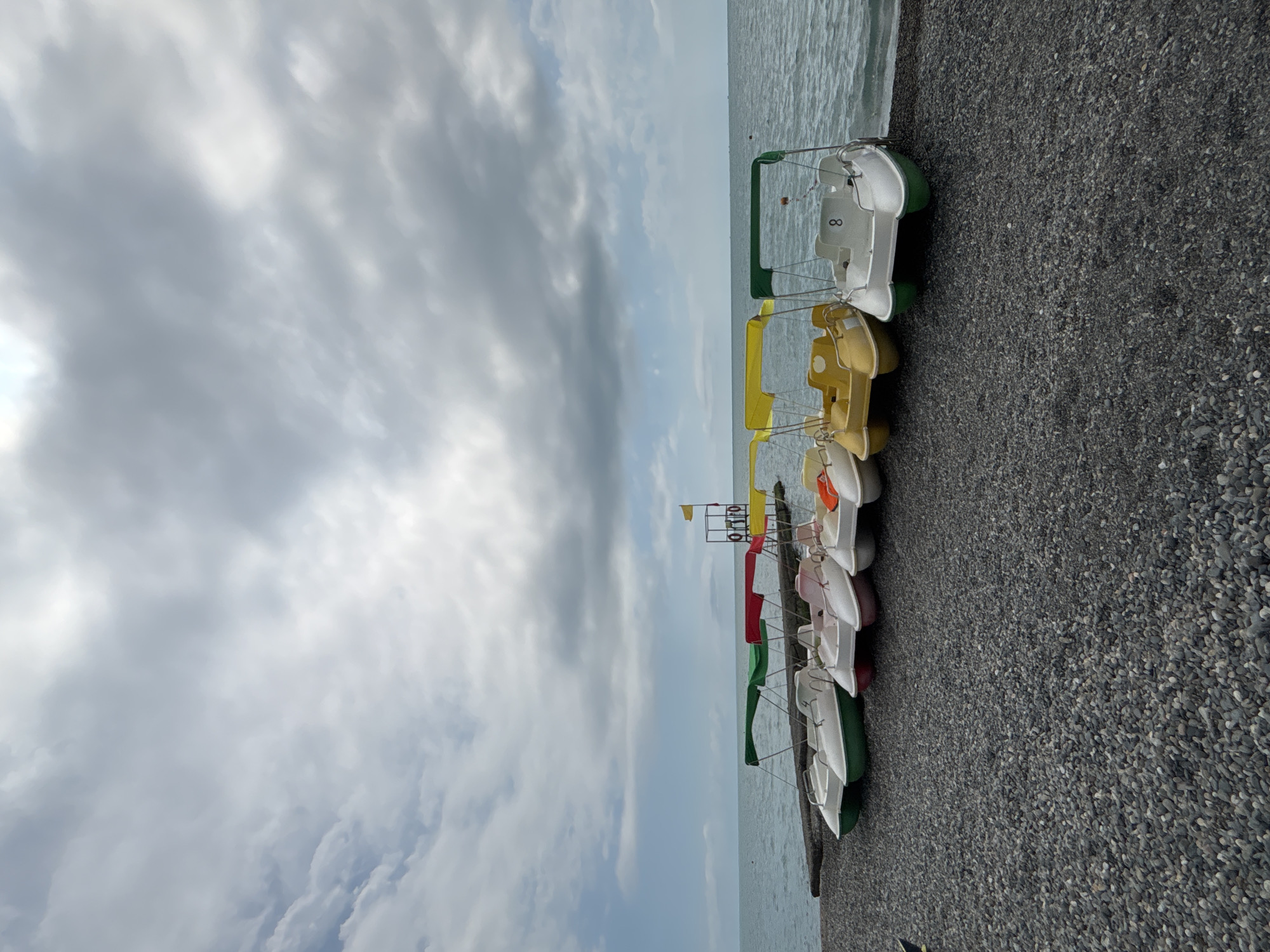 This is a vertical, eye-level shot of a row of pedal boats lined up on a pebble beach next to the sea. The sky is overcast with a mix of grey and white clouds, suggesting a cool or changeable day. The sea is a calm, muted blue-grey. The pedal boats are of various colors, including white, yellow, green, and red, and have canopies that match their hull colors or are green. Some of the white boats have the number "8" visible on them.
In the background, there is a lifeguard tower with a yellow flag at its top and a life preserver attached to it. The scene appears to be a recreational area by the water, likely a beach or a marina, with the pedal boats ready for use. The overall mood is tranquil, with no people clearly visible, giving a sense of quiet anticipation or the end of a busy period. The location context of Sochi, Russia, suggests this could be a beach along the Black Sea coast. The lighting indicates it is daytime, though the cloudy conditions diffuse the sunlight.