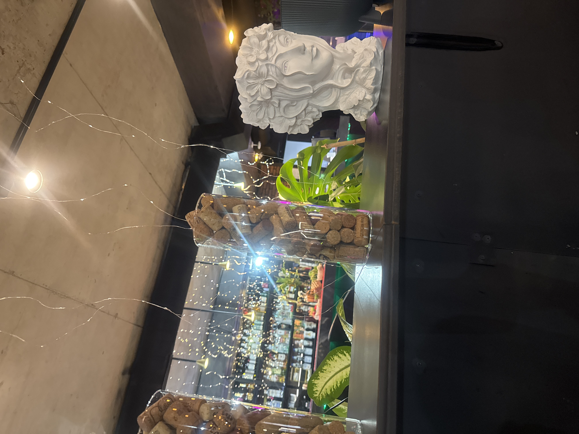 The image shows an interior setting, likely in a commercial space. A white bust sculpture of a woman with floral details in her hair is prominently displayed. Below the bust, a clear rectangular container filled with wine corks sits on a surface alongside leafy green plants, possibly Monstera. The backdrop is a blurred view of a liquor store, featuring illuminated shelves stocked with bottles. Soft fairy lights add a decorative element. While not explicitly stated, the description references the city of Tbilisi, Georgia, where the photo could have been taken.