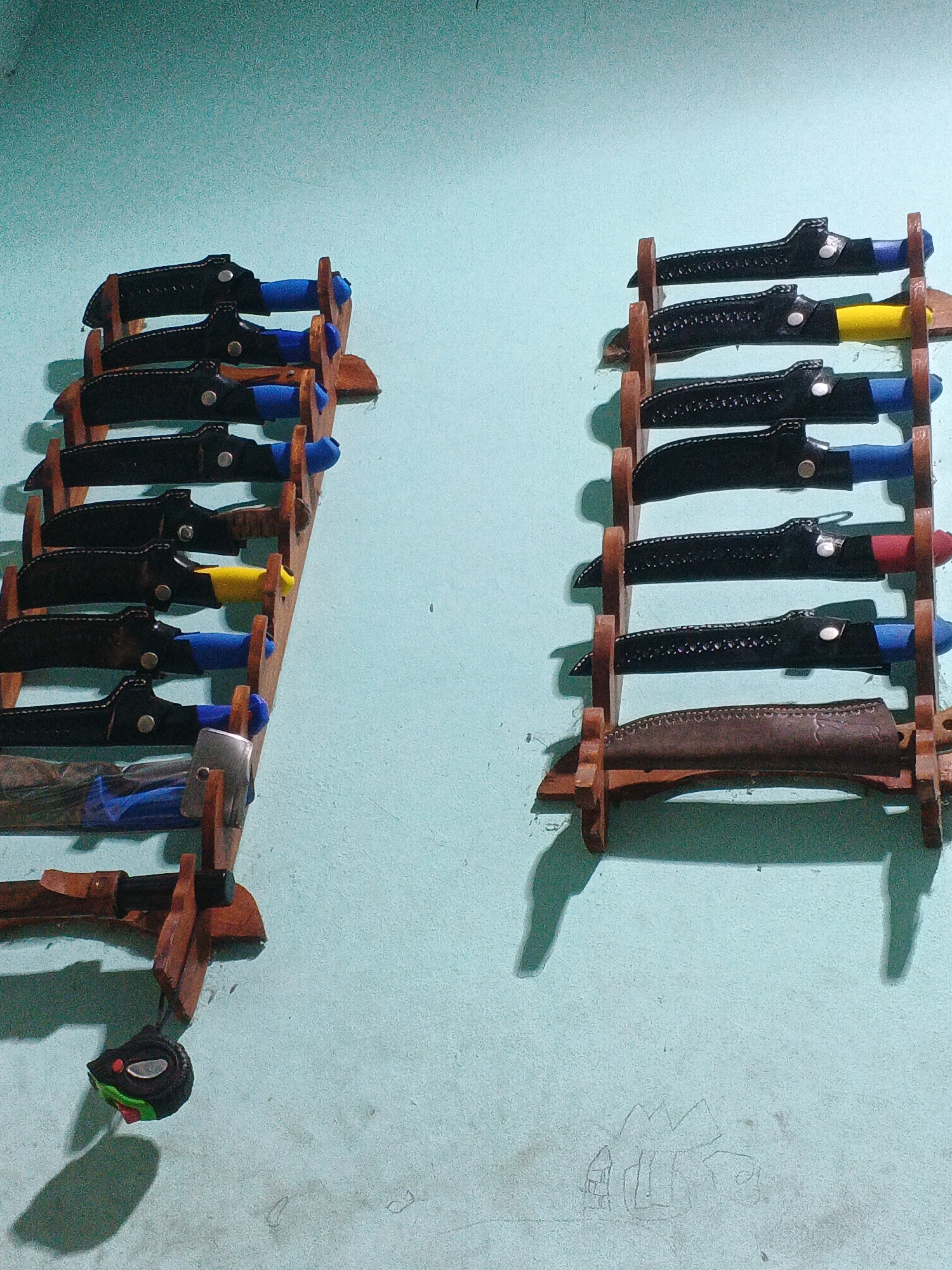The image depicts a wall in Kadu, Indonesia, adorned with two wooden knife racks holding several knives. The wall is painted a light blue color and shows some signs of age and wear. The knife racks are made of light-colored wood and are mounted vertically.
The left rack holds five knives, each with a blue handle and a black sheath. One of the knives on this rack also has a yellow portion on its handle.
The right rack holds five knives. The top four knives have black sheaths, and their handles are visible in varying colors including blue and yellow. The bottom knife on this rack has a brown leather sheath and a visible handle.
In the lower left corner of the image, a small, dark object with a metallic face is hanging from the left knife rack. This object appears to be a small tape measure. The shadows cast by the knives and racks suggest the lighting source is from the left. Some faint pencil markings resembling houses are visible on the wall in the lower right portion of the image.