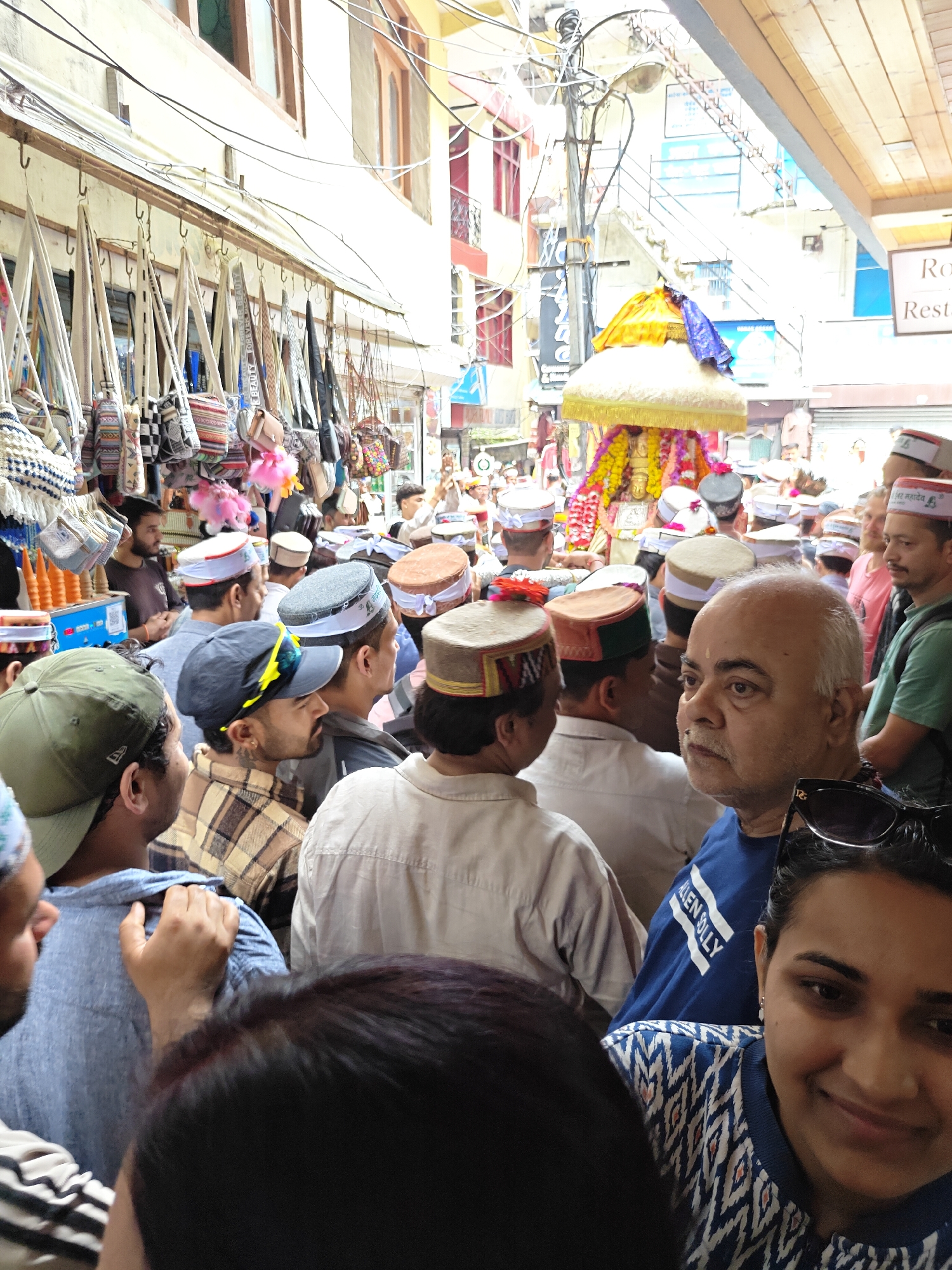This vibrant image captures a crowded street scene in Kasol, India, likely during a religious procession or festival. A dense gathering of people, mostly men, fills the narrow thoroughfare. Many individuals in the crowd are wearing traditional Himachali caps, distinctive for their round shape and colorful patterned bands. Some of these caps have visible text, such as "JAI MAHADEV" in Devanagari script, indicating devotion to Lord Shiva.
At the center of the crowd, a deity or idol is being carried, richly adorned with yellow fabric and garlands of colorful flowers, suggesting a significant religious event. On the left side of the street, a market stall is visible, displaying numerous bags and purses hanging from hooks. Some of these bags feature text like "BEAUTY FOREVER BEAUTY."
The scene is well-lit, indicating it is daytime. Overhead, a tangle of electrical wires crisscrosses between the buildings that line the street. In the foreground, a woman smiles towards the camera, while next to her, a man wears a blue t-shirt with "ALLEN SOLLY" printed on it. Another man, closer to the camera, has sunglasses pushed up on his head. The atmosphere appears lively and celebratory, characteristic of a local festival or gathering in a Himalayan town. Partially visible text in the upper right corner might spell "Restaurant."