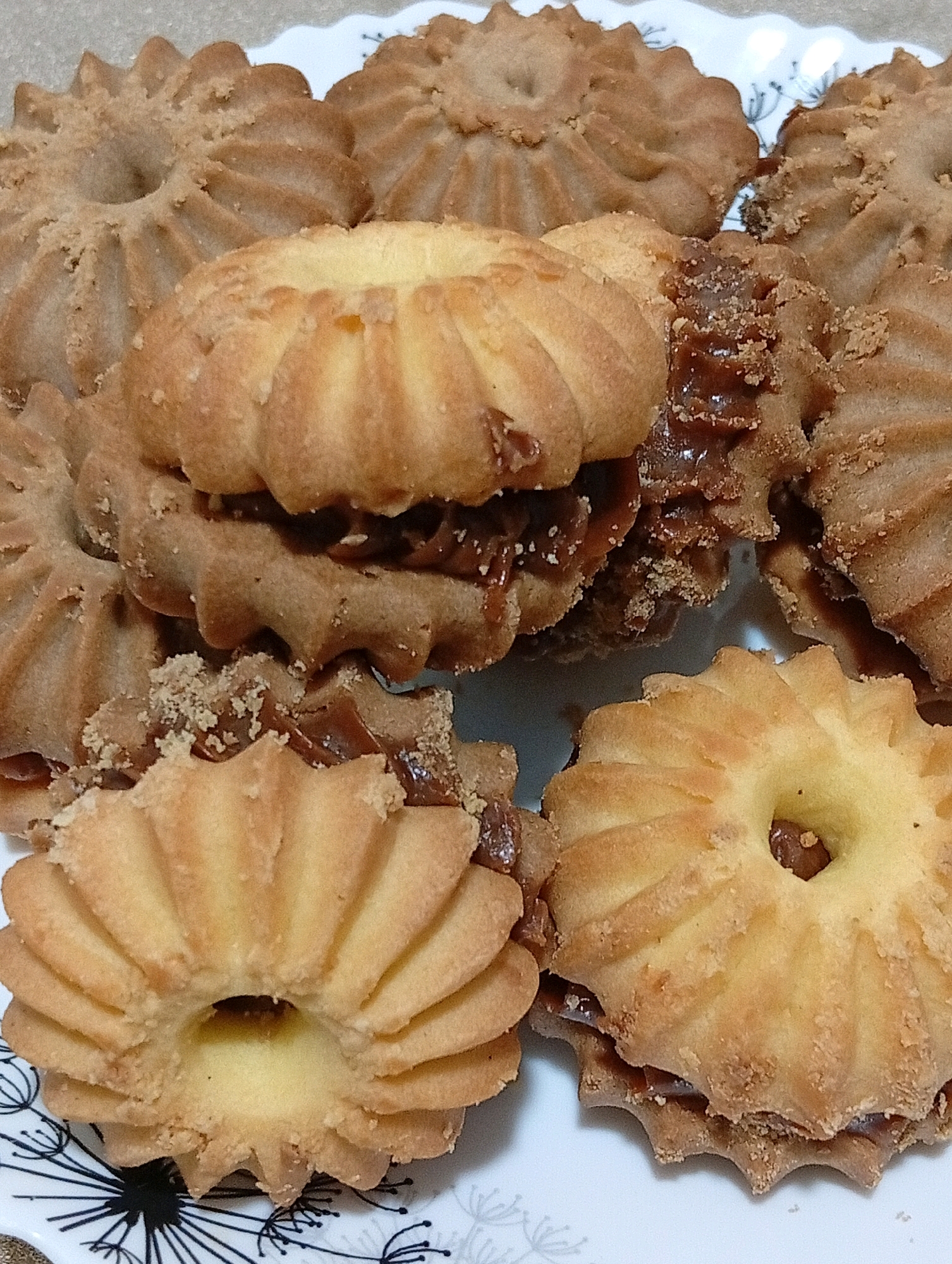 A close-up shot shows a pile of baked goods on a white plate with a black floral pattern. The baked goods appear to be sandwich cookies with a filling. They are arranged in a somewhat haphazard manner, with some cookies stacked on top of others. The cookies themselves are light brown and have a fluted edge. The filling visible between the cookies is a dark, glossy substance that appears to be caramel or chocolate. Some of the cookies have a dusting of crumbs on them. The lighting is even, and the focus is sharp on the cookies. The background is out of focus, but appears to be a light-colored surface.
