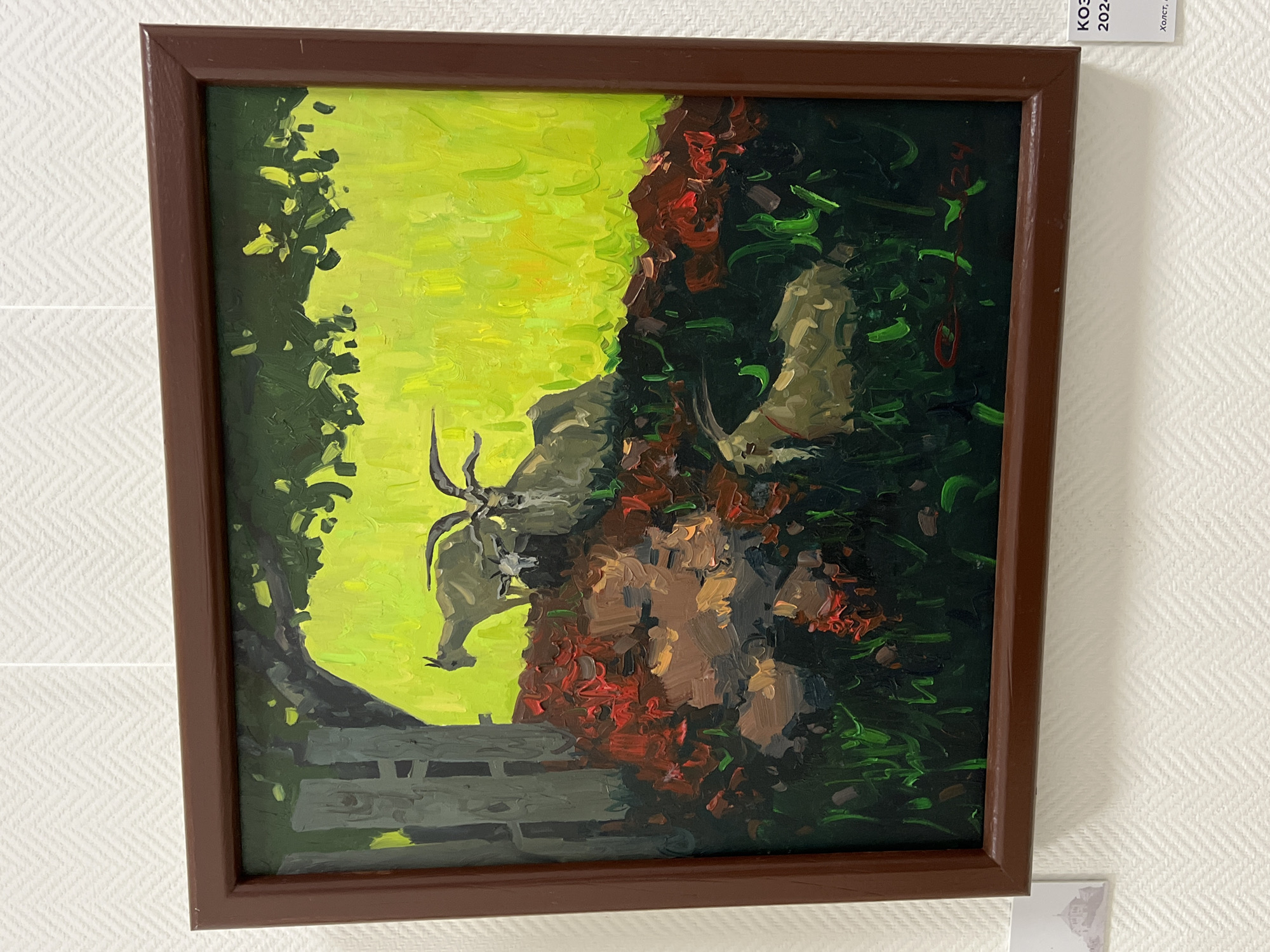 The image shows a framed painting of goats in a grassy area. The painting is oil on canvas, and the artist's signature and date are visible in the lower right corner, reading "КОЗЫ 2024". The scene depicts three goats, one lying down in the foreground and two standing behind it. The background is a bright yellow-green field under a tree. A wooden fence is visible on the left side of the painting. The painting is displayed on a white, textured wall, with a label below it containing the text "КОЗЫ" and "2024" and further text in Russian. There is also a small print of a building in the bottom left corner.