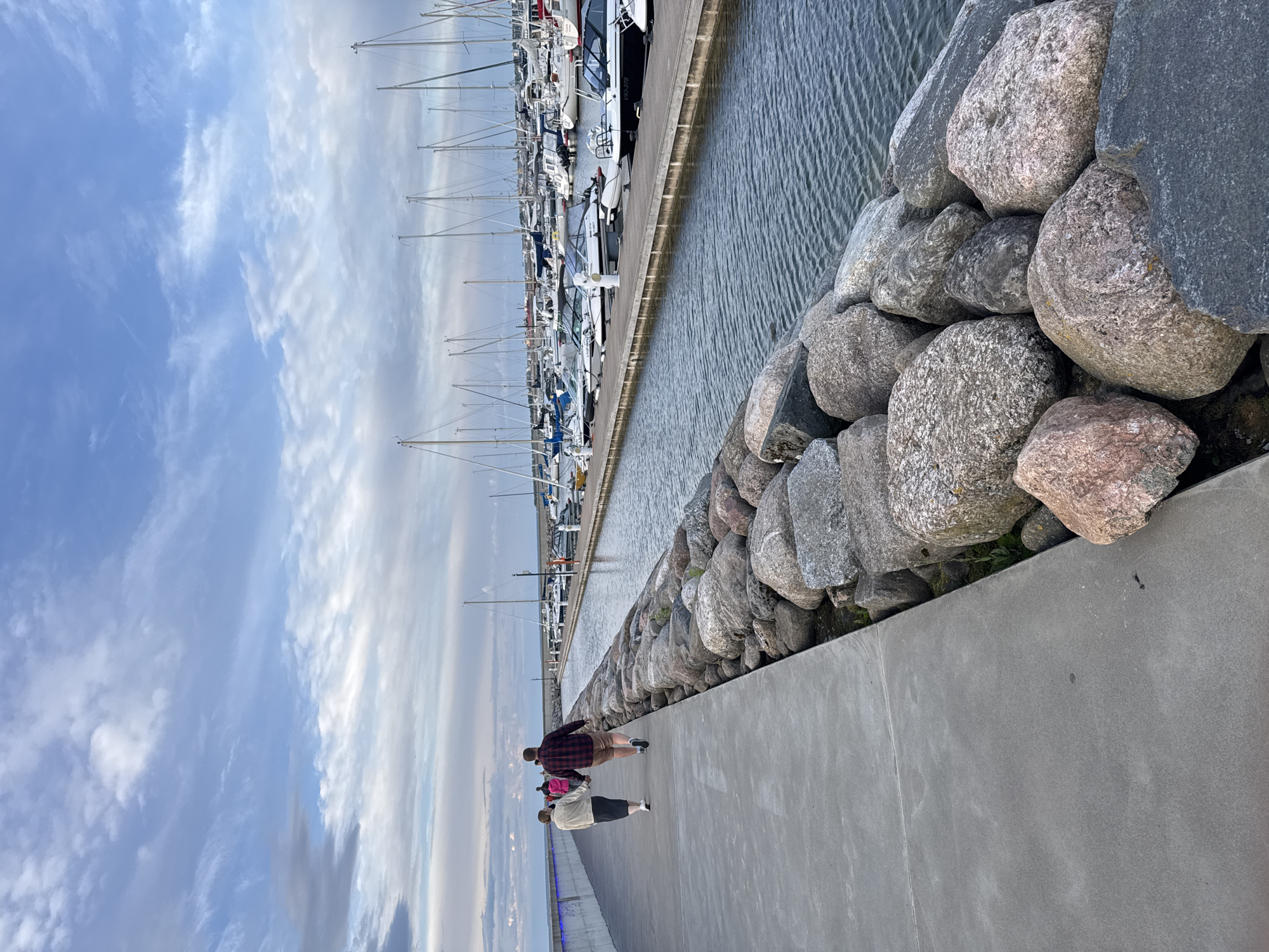 This is a vertical, eye-level shot of a promenade and a marina on a cloudy day. The promenade is on the left, made of grey concrete. It is bordered on the right by a rock seawall, constructed of large, grey and pinkish boulders. Beyond the rock seawall is a canal filled with dark, rippling water. On the other side of the canal is a marina, with numerous sailboats and motorboats docked. The boats are white, grey, and black. Many have tall masts. The sky above is blue with scattered, wispy white clouds. In the foreground on the promenade, a man in a red and black plaid shirt and beige shorts walks hand-in-hand with a woman wearing a light-colored jacket and a dark skirt. Behind them, further down the promenade, two more people are walking. The location is likely the Old City Marina or the adjacent Pirita Marina, both located in Tallinn, Estonia, given the architecture and the presence of a marina in a Baltic city.