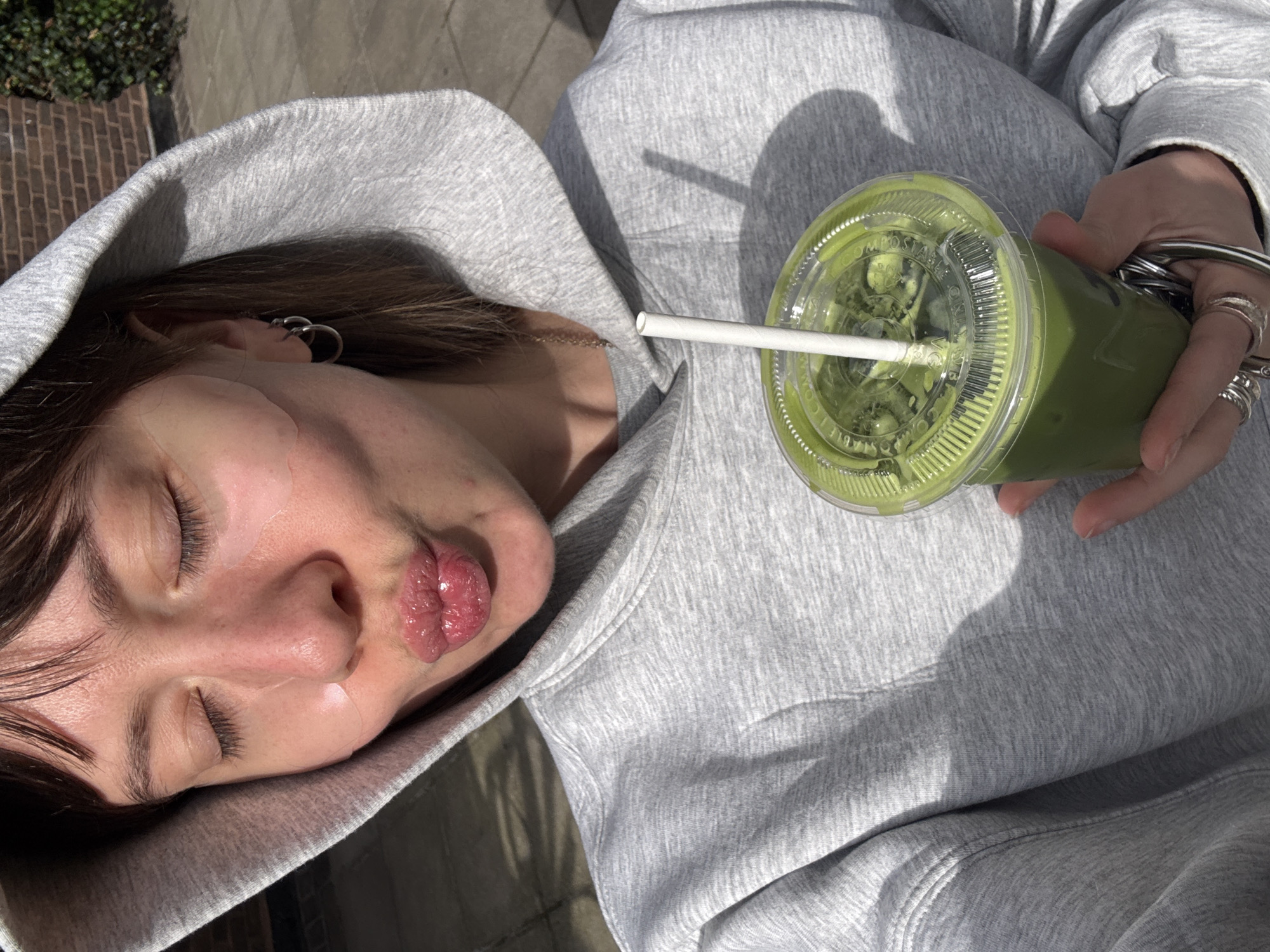 A close-up, rotated image captures a person outdoors in what appears to be London, United Kingdom, under bright, sunny daylight. The subject, visible from a high angle, wears a light gray hooded sweatshirt with the hood up. Their eyes are closed, and their lips are distinctly pursed into a pout. Several light-colored patches, possibly hydrocolloid or similar skincare products, are visible on their cheek and forehead area. Dark hair frames their face, and the visible ear is adorned with multiple silver hoop earrings. A thin chain is also visible near their neck.
The person's right hand, decorated with several silver rings, holds a clear plastic cup containing a vibrant green beverage, strongly resembling a matcha latte. A white paper straw extends from the cup's transparent lid. Faint text is visible on the lid, including the words "COMPOSTABLE" and "STABLE". Strong sunlight casts crisp shadows across the person's clothing and the drink, emphasizing the bright outdoor conditions. The blurred background suggests an urban setting, with hints of brickwork and green foliage. The overall scene conveys a casual, relaxed, and possibly playful moment.