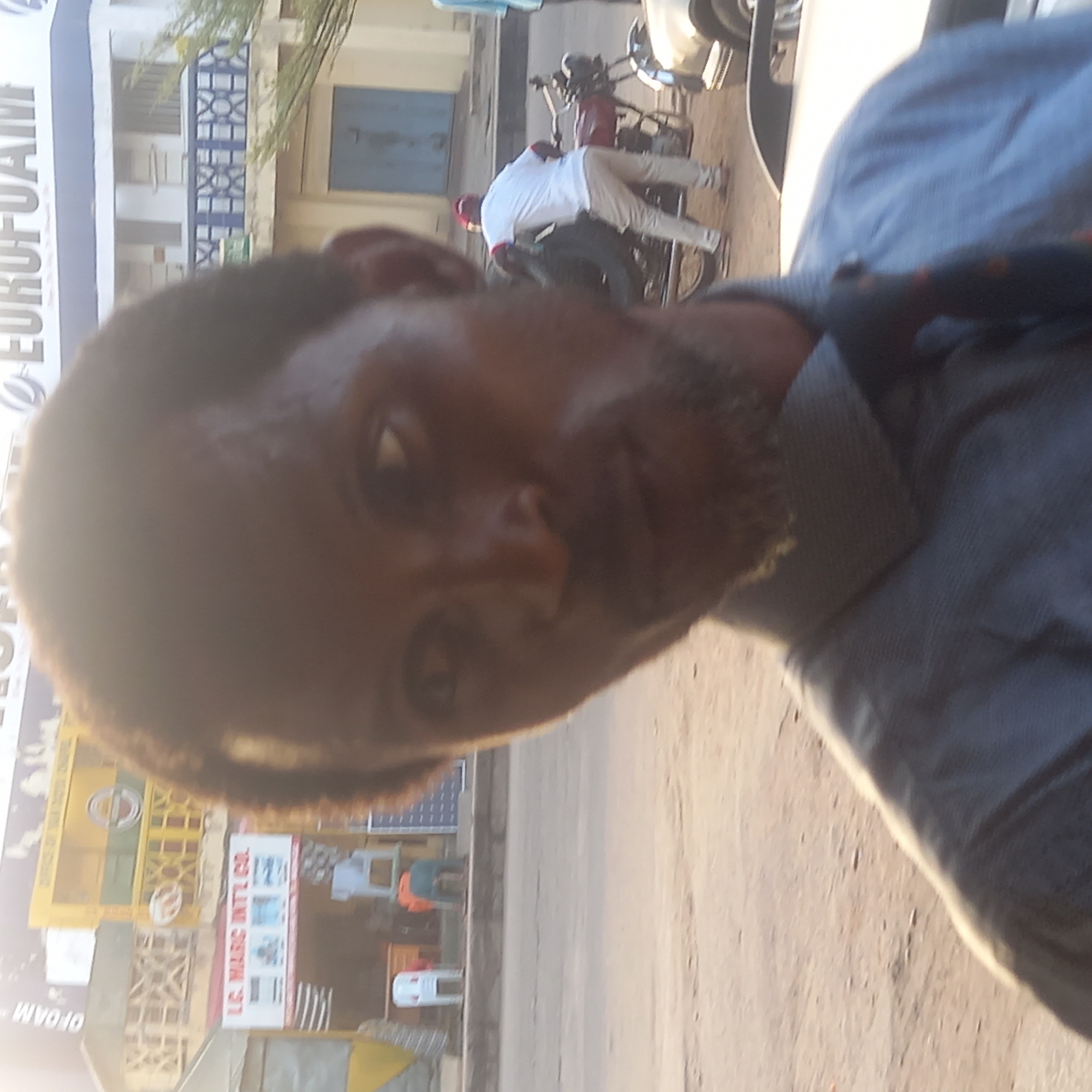A man wearing a grey collared shirt is taking a selfie in front of a building in what appears to be Sardauna, Nigeria. The building has signs for "EUROFOAM" and "L.C. MARC INTL CO." A man in white sits on a motorcycle in the background.