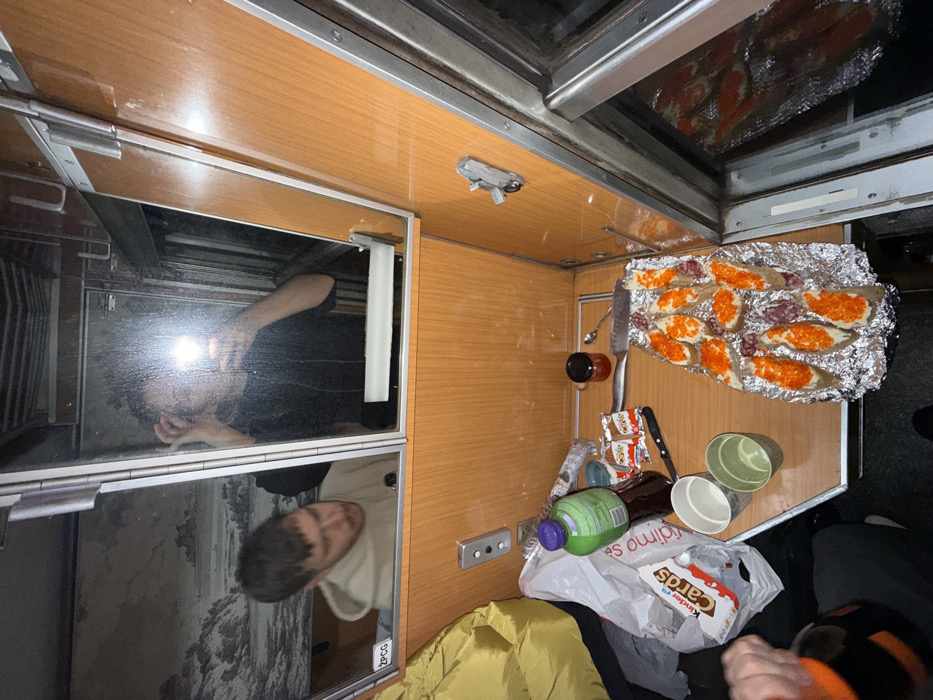 An overhead shot displays a messy train compartment table. Caviar-topped bread sits on foil, alongside condiments, cups, and wrappers. A Kinder box and a green bottle are visible amongst the clutter. The table is wood-paneled. Mirror cabinets reflect two people; one holding a light, the other in a light sweater.