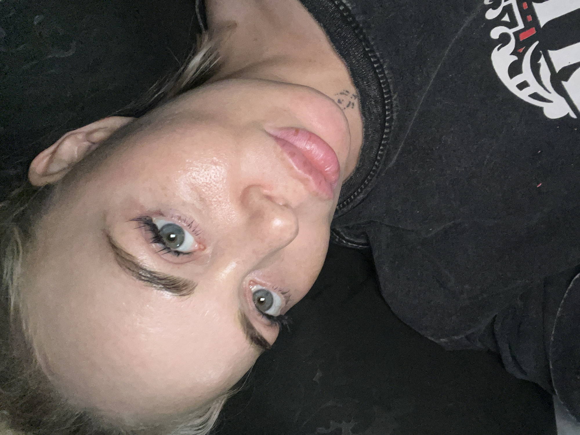 A close-up, high-angle, indoor selfie shows a woman with blonde hair and blue eyes. She is looking at the camera, her head tilted to the right. The woman is wearing a dark t-shirt with a graphic design on the front. Her makeup is visible, including mascara and lip gloss. The background is dark and appears to be a room with soft lighting.