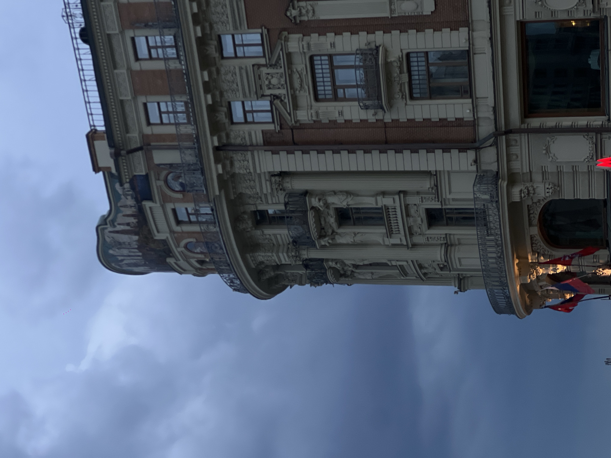 The image shows a multi-story building with ornate architectural details. The building has a curved corner, visible windows with iron balconies, and detailed cornices. Some red flags are hanging from the building. The sky in the background is overcast and a dark grey color. The location of the building is unspecified.