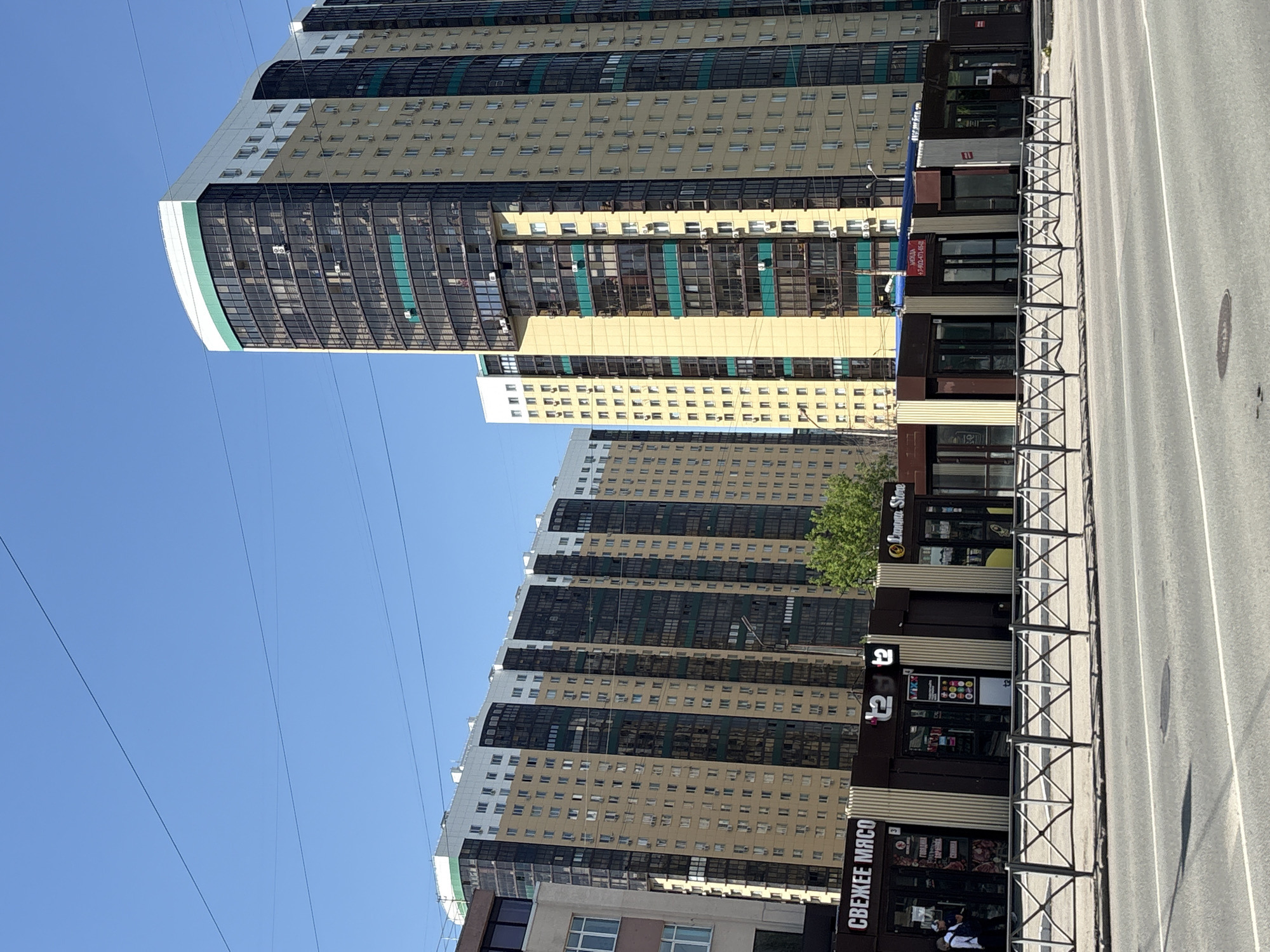 The image depicts a street scene in Perm, Russia, showcasing modern architecture and local businesses. Several high-rise apartment buildings dominate the background, characterized by a mix of light brown and dark reflective glass facades. These buildings feature numerous windows and what appear to be glass-enclosed balconies.
Along the street level, several retail establishments are visible. One notable business has signage written in Russian, including the phrase "СВЕЖЕЕ МЯСО" (Fresh Meat). A person is standing near this store.
A metal fence runs along the edge of the sidewalk, separating it from the street. The street is asphalt and shows typical markings of a roadway.
The sky is clear and blue, suggesting a sunny day. Overhead power lines stretch across the scene.