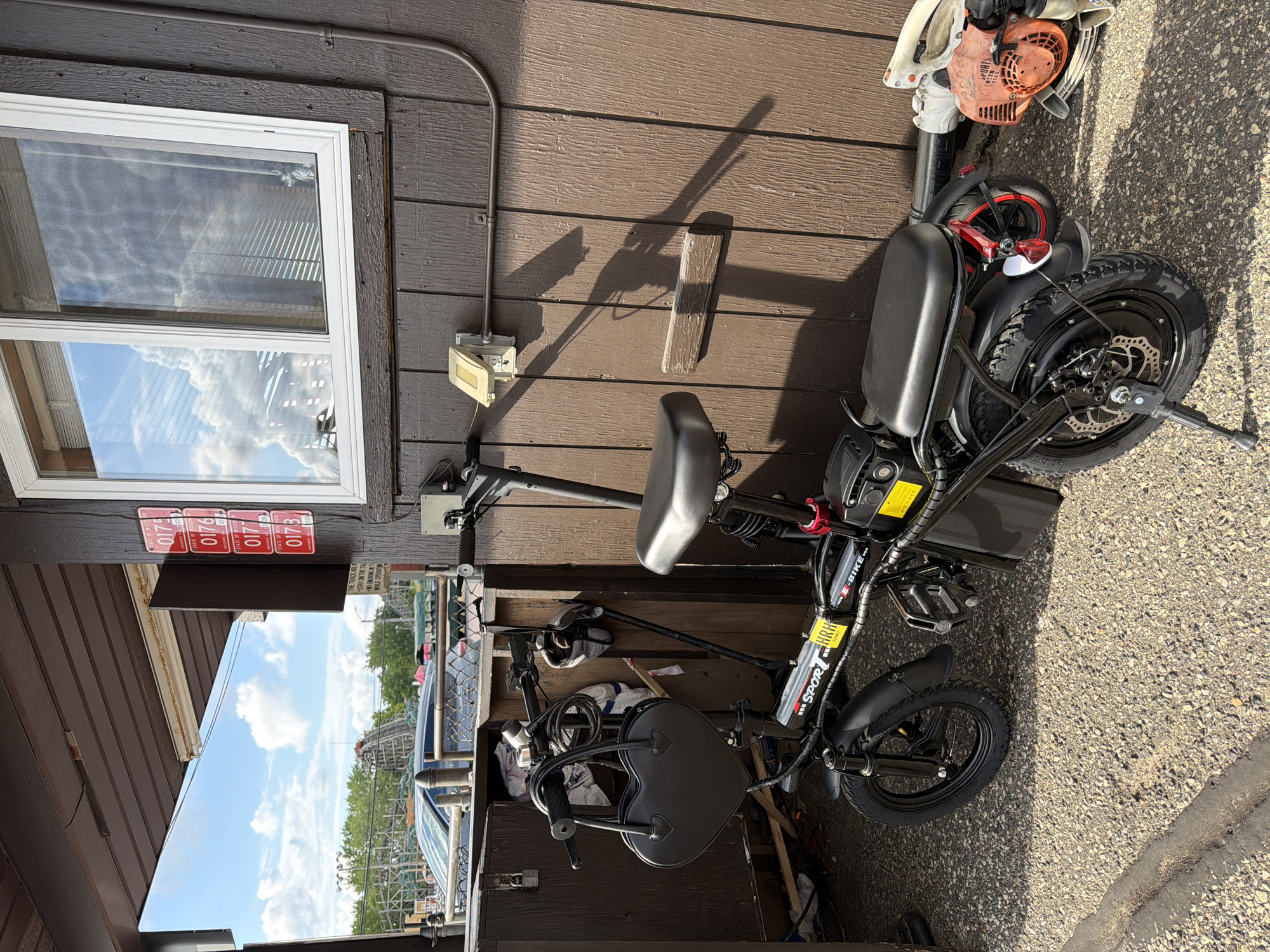 The image displays an outdoor scene featuring a black folding e-bike parked in front of a brown wooden building, with an amusement park visible in the background under a blue sky with white clouds. The location is the city of Lake Delton, United States of America, confirmed by "STATE OF WISCONSIN" text on several red rectangular tags affixed to the building.
The e-bike, positioned centrally, is black with "SPORT" and "E-BIKE" branding on its frame. It features two black padded seats, small black wheels with thick tires, and visible disc brakes on the rear wheel. A kickstand is deployed on the rear left. A black heart-shaped handbag hangs from the handlebars. A yellow warning label is present on the battery pack.
The building on the right has brown, vertically-paneled wooden siding. A white-framed window on the upper right reflects the sky and clouds, with interior blinds partially visible. Below the window, a light-colored electrical outlet with a yellow-tan cover is mounted, and a grey metallic conduit runs horizontally and then vertically along the wall. Four red rectangular tags, stacked vertically, are affixed to a dark wooden pillar on the building's facade. Each tag reads "STATE OF WISCONSIN" and is numbered sequentially: "0175," "0176," "0177," and "0173" (out of sequence at the bottom). A small, weathered wooden block is also attached to the siding below the conduit.
Through an open section to the left of the building structure, a large roller coaster with light-colored tracks and support beams is clearly visible. Green trees are abundant, and a grey car is partially visible behind a metal railing or fence. The ground is a light-colored gravel or coarse asphalt surface. Long shadows cast by the e-bike and the building indicate direct sunlight from a relatively low angle. To the right of the e-bike's rear wheel, a red and white gasoline-powered leaf blower rests on the ground. No individuals are present in the scene.