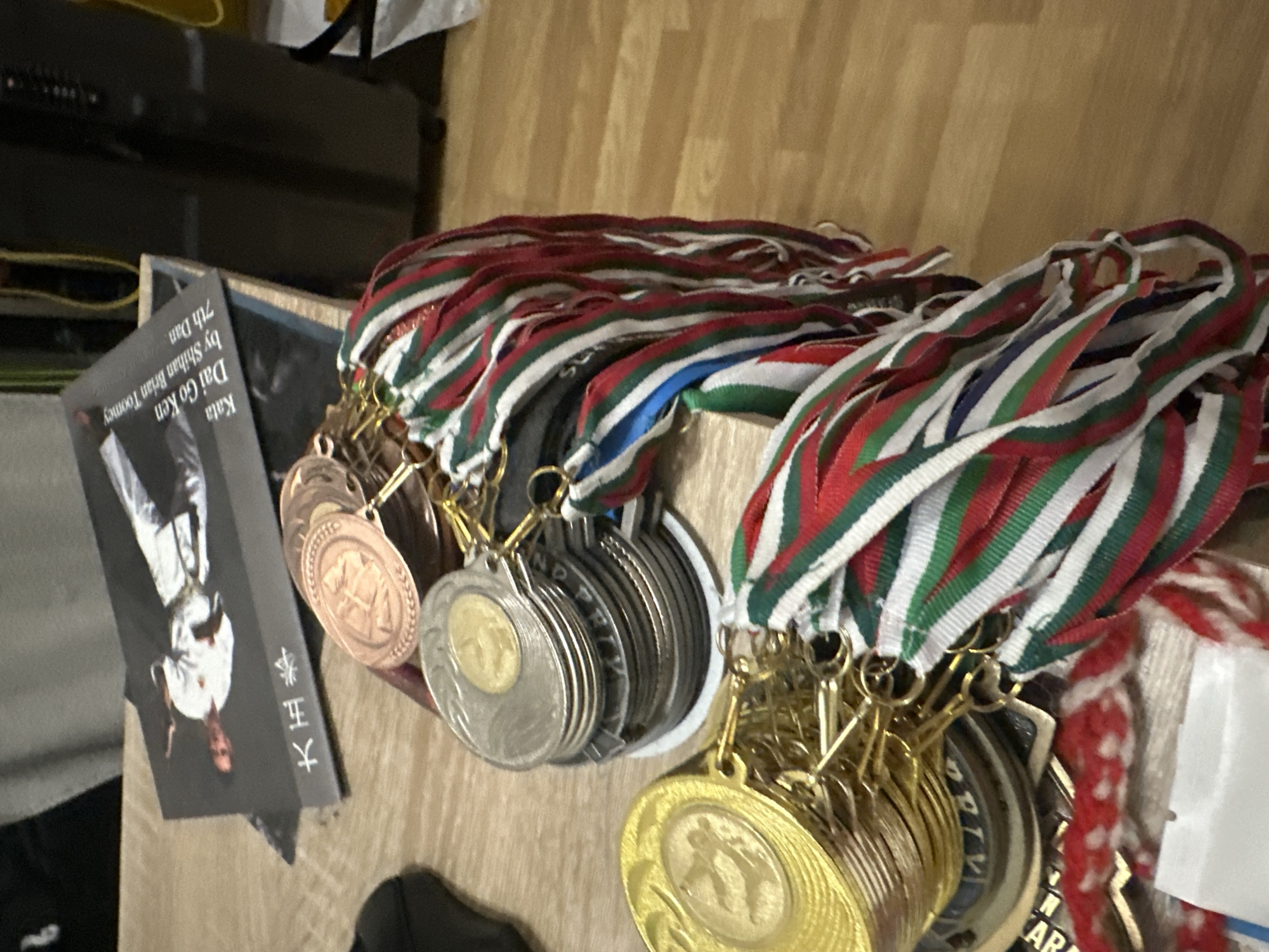 The image depicts an indoor scene in the city of Unknown, Unknown, primarily focused on a collection of martial arts medals and a book resting on a light-colored wooden table with visible grain.
On the left, a substantial stack of gold-colored medals is present, with the topmost medal displaying an embossed figure in a martial arts stance. Adjacent to these and slightly to the right, a stack of silver-colored medals with gold-colored centers is observed. Behind these, a stack of bronze-colored medals is partially visible. All medals are attached to numerous striped ribbons, predominantly red, green, and white. Some ribbons also feature a blue stripe. The ribbons are secured to the medals with small, gold-colored clasps.
Behind the stacks of medals, a dark-covered book or pamphlet is placed diagonally on the table. Its cover features an image of a person in a white martial arts uniform (gi) executing a dynamic pose. Visible English text on the cover, oriented vertically, reads "Kata Dai Go Ken by Shihan Brian Toomey 7th Dan." To the left of the image, two large Chinese characters, "大正" (Taisho), are also present.
The background includes a dark, indistinct object, possibly furniture or equipment, positioned on the right. Yellow cables are visible near this object, and a crumpled white item, potentially a bag, is on the far right. The scene is illuminated, casting minor reflections on the metallic surfaces of the medals.