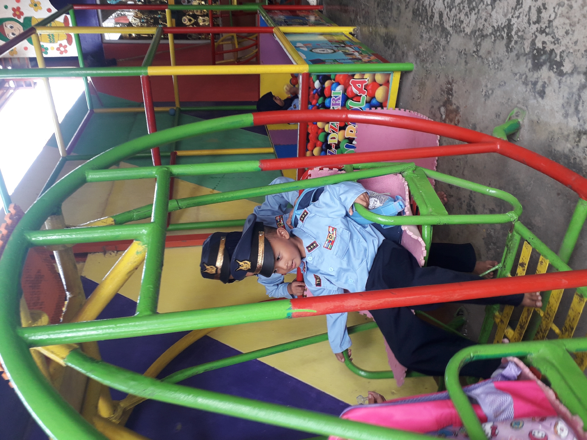 In Cankuang, Indonesia, two young boys in light blue uniforms and dark caps sit on a green swing set with red and yellow accents. The boy in the foreground faces right, looking away from the camera, while the boy behind him faces left. Both are seated on colorful cushions. The background features a ball pit filled with multi-colored balls, surrounded by yellow and green structures. The ground is a wet, mottled grey concrete.
