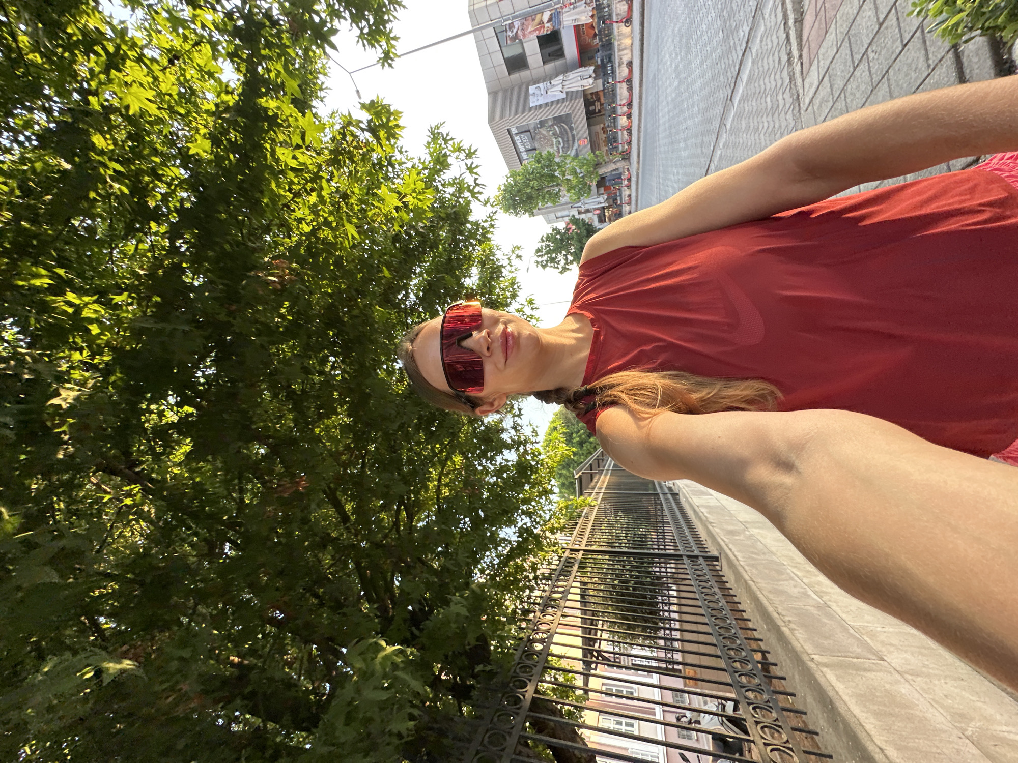 In Fethiye, Turkey, a woman with blonde hair braided down her back and red mirrored sunglasses stands on a sidewalk beneath a large tree. She is wearing a sleeveless red t-shirt with a Nike logo. To her left is a black metal fence with an ornate design, behind which is a building with a light-colored facade and windows. The sidewalk is paved with cobblestones. Across the street, there are shops and buildings, one with a large advertisement for a burger restaurant. Several red electric scooters are parked in a row along the sidewalk. The lighting suggests it is late afternoon or early evening.
