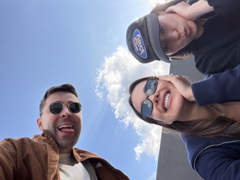 A low-angle photograph captures three individuals outdoors against a clear blue sky with scattered white cumulus clouds, in what is presumed to be Tel-Aviv, Israel. In the upper left quadrant, a male subject, wearing gold-rimmed sunglasses and a brown corduroy jacket over a light shirt, has his mouth open with his tongue extended, directing his gaze towards the camera. In the lower left quadrant, a female subject with long dark hair, wearing tortoise-shell sunglasses and a dark blue zippered hoodie, smiles broadly with her left hand resting on her left cheek, also looking towards the camera. Adjacent to her on the right, in the lower right quadrant, a second female subject wears a dark baseball cap with a blue oval "Ford" logo and a dark zippered jacket; she has pursed lips and her right hand resting on her right cheek, with her gaze directed towards the camera. A dark, non-descript horizontal barrier or wall is visible in the background, beneath the sky.