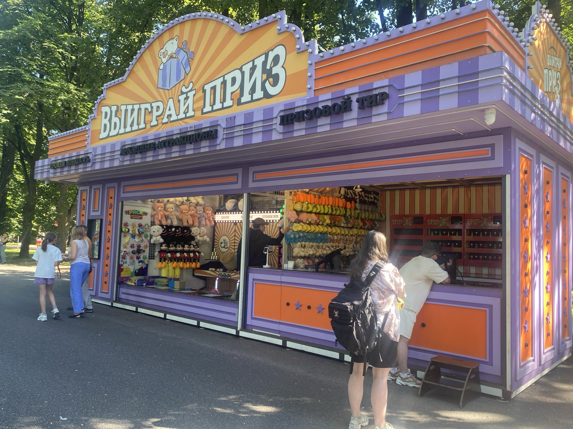 The image depicts an outdoor carnival game booth in Saint Petersburg, Russia. The booth has a vibrant, colorful facade with purple and orange stripes and a large sign in Russian that reads "ВЫИГРАЙ ПРИЗ" (Win a Prize). To the right of this sign, another sign reads "ПРИЗОВОЙ ТИР" (Prize Shooting Gallery). The booth features large display windows showcasing stuffed animals as prizes, including teddy bears and Mickey Mouse characters. There are also shelves stocked with various colorful plush toys. Several people are interacting with the booth. On the left, two young women are standing near the display window. In the center of the booth, an employee is handing a toy to a customer. On the right, a man is aiming a rifle at targets inside the booth, with another person observing. The scene is set against a backdrop of lush green trees, suggesting a park or outdoor entertainment area. The ground is paved asphalt.