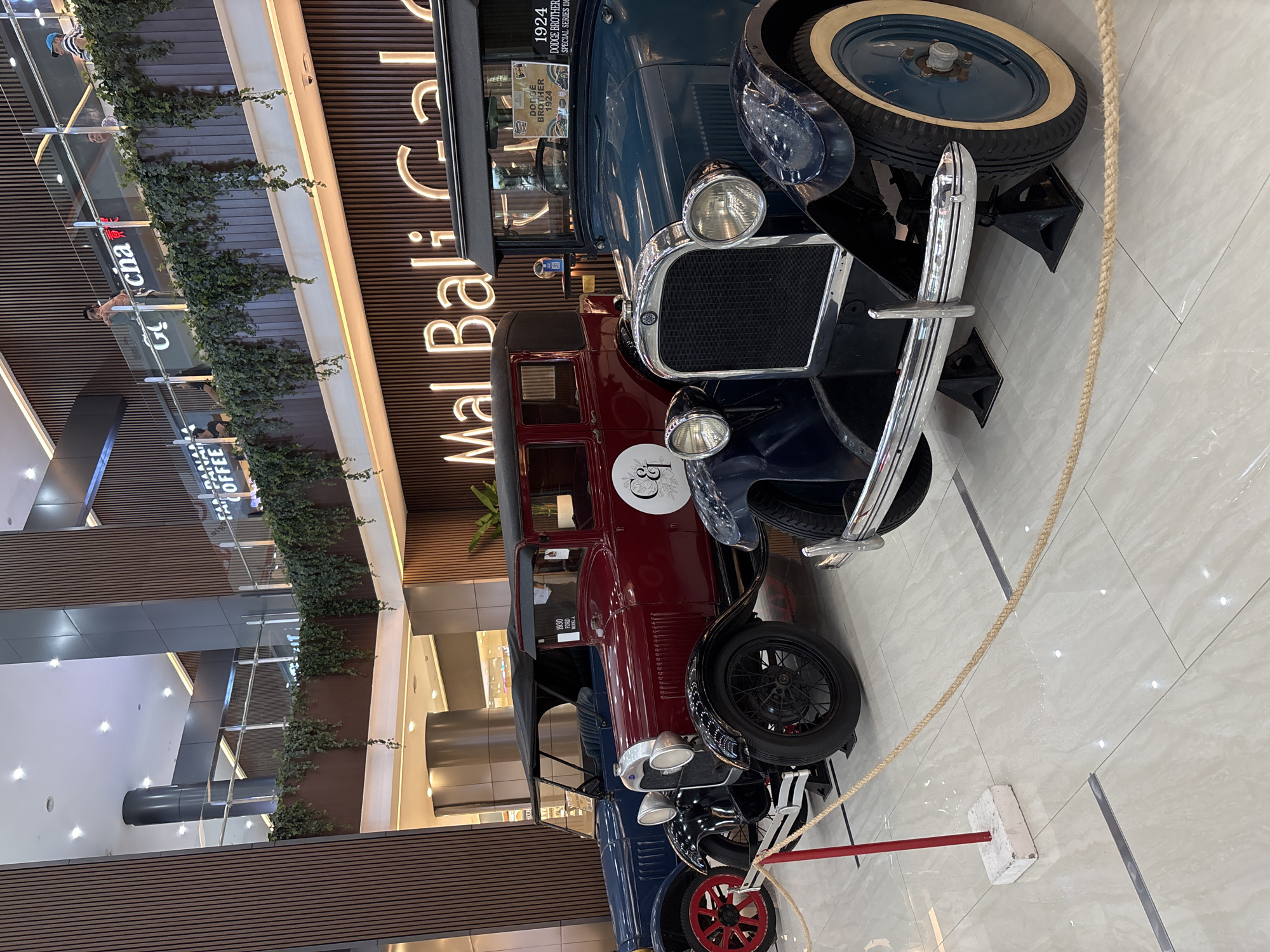 Here is a description of the image:
The image shows a display of vintage cars inside a shopping mall. Specifically, a Dodge Brothers Special Series 116 from 1924, and a Ford Model A from 1930. These cars are roped off, positioned in front of a "Mal Bali Galeria" sign, within a contemporary mall environment. The mall also houses a "Gcna COFFEE" shop. The picture suggests this display is likely within a shopping center in Bali, Kuta, Indonesia.