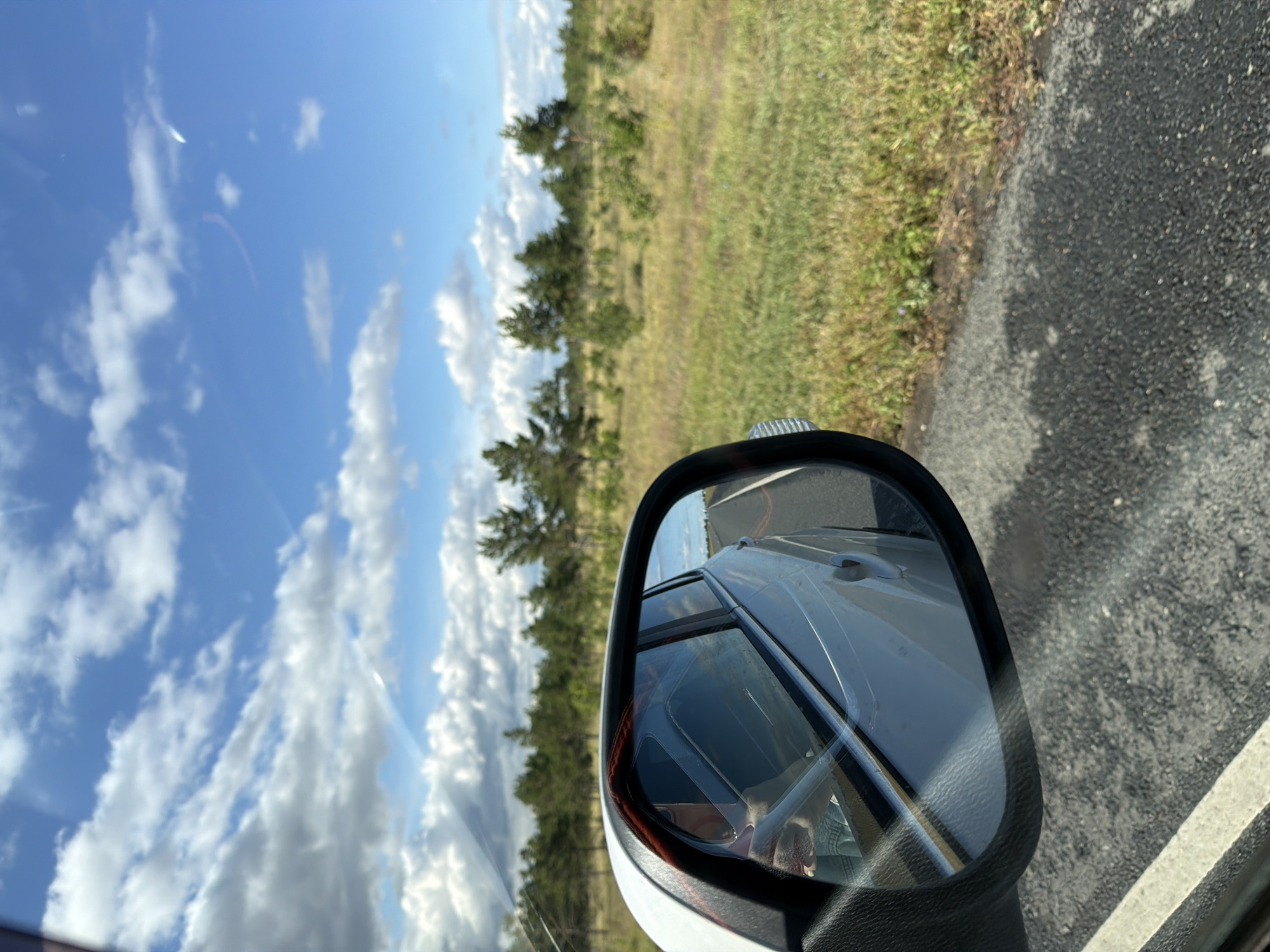 The image shows a reflection in a car's side mirror, showing the side of a white vehicle traveling on a road. The reflection also captures a view of the landscape, which includes a field with dry grass, scattered trees, and a blue sky with scattered clouds. The image's angle suggests it was taken from inside the car, looking out the window towards the side mirror. There are no people or specific landmarks visible in the reflection that would indicate a location in Tavolzhanka, Russia. The scene is a general depiction of a roadside landscape.