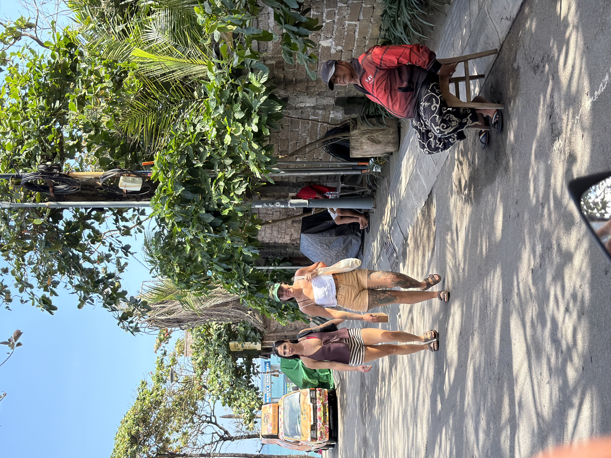 This image captures a street scene in North Kuta, Indonesia, likely during the daytime under sunny weather, as evidenced by the strong shadows cast on the ground. In the foreground, two young women are walking down a paved street lined with lush green foliage. The woman in front wears a dark brown top with thin straps and striped shorts, while the woman behind her has on a white tank top and tan shorts. Both are wearing sandals. Their attire suggests a warm climate and a casual atmosphere.
Further up the street, a person is seated on a small wooden stool, wearing a red and black jacket and a dark patterned skirt. They appear to be observing the surroundings. Behind them, a brick wall and some assorted items, including what looks like a wooden crate and some electrical wiring, suggest a residential or commercial area.
A small, brightly decorated van with the text "BALI FRUIT NINJA" visible on its side is parked on the left side of the street. The van is orange with colorful floral patterns. The overall scene is one of everyday life unfolding in a tropical urban environment. The presence of trees, the style of the van, and the clothing of the people provide strong contextual clues to the location. The lighting suggests it is likely morning or afternoon.