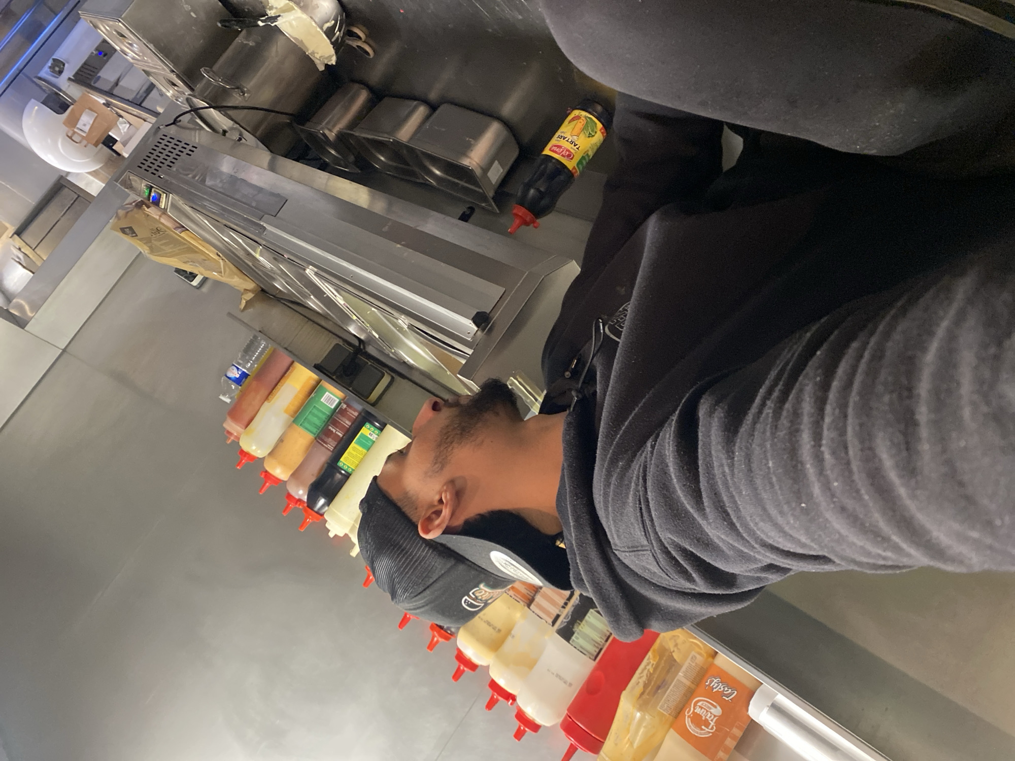 The image depicts a commercial kitchen or food service area in Schiltigheim, France, captured from a low angle, looking upwards and across the counter.
A male individual is visible in the left foreground, positioned with his back partially to the camera, looking towards the right side of the frame. He is wearing a black hooded sweatshirt and a black baseball cap, possibly reversed. He has a short beard.
To the individual's left, on a light-colored counter or shelf, a linear arrangement of approximately ten plastic squeeze bottles with red caps is present. These bottles contain various sauces, discernible by their colors as red, white, yellow, light brown, dark brown, and black. Another bottle, labeled "Maison Tartare" with a yellow label and a red cap, is visible on a lower dark surface to the right of the individual.
The background primarily consists of industrial stainless steel equipment, including a food preparation counter or bain-marie with several rectangular stainless steel inserts. Above this counter, additional stainless steel elements are visible, along with a white plastic container and a brown paper bag on an elevated shelf. A blue plastic water bottle is also present further in the background. The surrounding walls appear to be a light grey or stainless steel, consistent with a professional kitchen environment. No direct interactions or specific actions beyond observation are depicted for the individual.