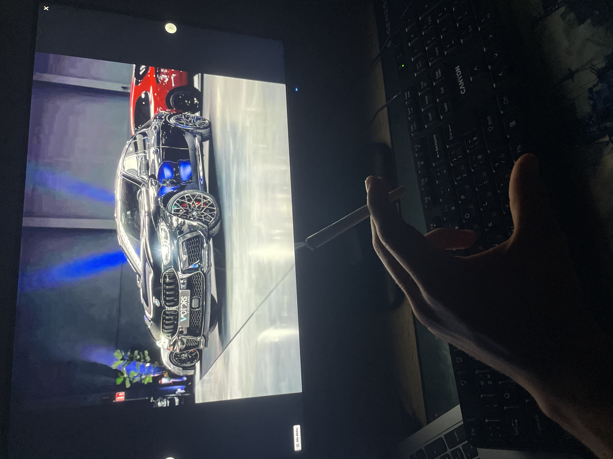 The image depicts an unknown indoor setting, dimly lit. A dark-colored computer monitor occupies the upper two-thirds of the frame, displaying a digital image of three luxury automobiles. The central vehicle is a highly reflective black BMW M8 Gran Coupe with custom multi-spoke wheels, exhibiting blue reflections on its bodywork and a front plate-like element that reads "MOTORS premium cars." A silver vehicle is partially visible to the left, and a red vehicle is partially visible to the right, both positioned behind the BMW. The background of the displayed image features dark walls accented with vertical blue lighting and some green foliage. User interface elements from an image viewer are visible on the monitor's display: "Alle Bilder" (German for "All Pictures") in the lower-left corner, an 'x' icon in the upper-right corner, and a white right-arrow navigation icon on the right edge. A small blue LED indicator light is visible on the bottom bezel of the monitor.
In the lower portion of the image, a human hand is visible, positioned centrally. The hand is holding a thin, white cylindrical object between the index and middle fingers, with its tip glowing orange, consistent with a lit cigarette. To the right of the hand, a black "CANYON" branded computer keyboard is partially visible, displaying a green illuminated indicator light. A segment of a laptop computer is visible on the far left. All items rest on a wooden surface.