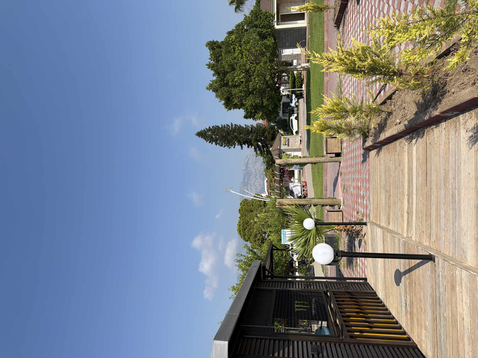 This image depicts an outdoor scene in Kemer, Turkey, on a clear day with a bright blue sky. In the foreground, a wooden walkway is illuminated by sunlight, casting shadows from two lampposts. To the left, a dark-colored, modern building with slatted walls and a metal fence encloses an interior visible through the fence, displaying a "MONSTER" logo. To the right of the walkway, a row of small, yellow-green coniferous trees are planted in a raised garden bed.
Further into the scene, a large palm frond is prominent, with two white globe-shaped lights on dark poles standing behind it. Beyond this, a pathway paved with red and white interlocking bricks leads towards a grassy area. In the middle ground, several trees with lush green foliage are scattered across the landscape. A tall, slender pine tree stands to the left of a wider, more mature tree on the right. Several buildings, including one with a gray brick exterior and an open doorway, are visible in the background. A white car is parked on a paved surface in front of some of these buildings. In the distance, a hazy mountain range is visible under the clear sky. The overall impression is that of a well-maintained public space or resort area.