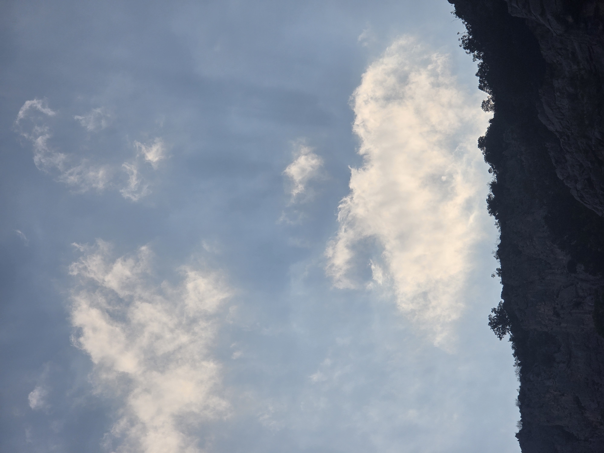 The image depicts a dramatic sky filled with luminous clouds, suggesting either sunrise or sunset. The clouds are varied in density, with some appearing wispy and translucent, while others are thick and illuminated from behind, casting soft rays of light. Below the sky, a silhouetted ridge of rugged, rocky terrain is visible, densely covered with dark vegetation and trees. The topography appears steep and uneven. The overall scene is one of natural grandeur, with the stark contrast between the illuminated sky and the darkened landscape creating a sense of depth and drama. This view could be from a vantage point overlooking the mountainous outskirts of Podgorica, Montenegro, possibly from the Gorica hill or a similar elevated area offering expansive sky and landscape views.