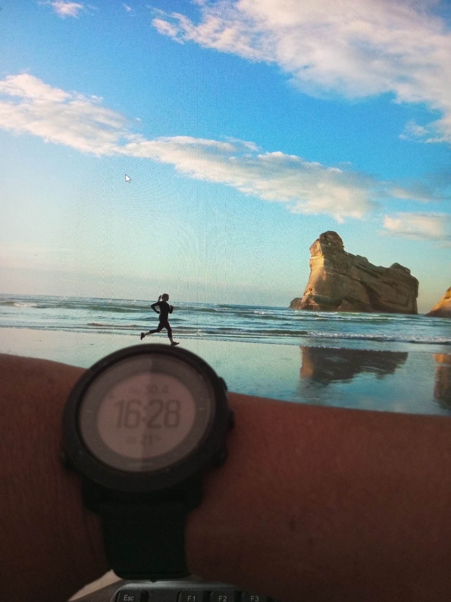 The image captures a first-person perspective, focusing on a person's left arm adorned with a black smartwatch, positioned over a computer keyboard, facing a computer monitor. The monitor displays a scenic desktop wallpaper of a beach at what appears to be late afternoon.
The wallpaper depicts a clear blue sky with scattered white, cumulus clouds. Below the horizon, a calm ocean shows gentle waves breaking onto a sandy beach, with a wet reflective surface near the waterline. A distinctive, large, light-brown rock formation, resembling a sea stack or cliff, dominates the right side of the screen, its reflection clearly visible in the water and wet sand. In the mid-ground, a dark silhouette of a person running along the water's edge is visible. A white mouse cursor is also present on the screen.
The smartwatch in the foreground prominently displays "16:28" as the time. Above it, smaller text reads "10:4", and below, "0.21", likely indicating a date or other metric. The watch features a dark casing and a black strap. The keys "Esc", "F1", "F2", and "F3" of a computer keyboard are partially visible at the bottom of the frame.
The photo combines an indoor setting (person viewing a screen) with the outdoor coastal environment displayed on the monitor. The photo was taken in Lhokseumawe, Indonesia.