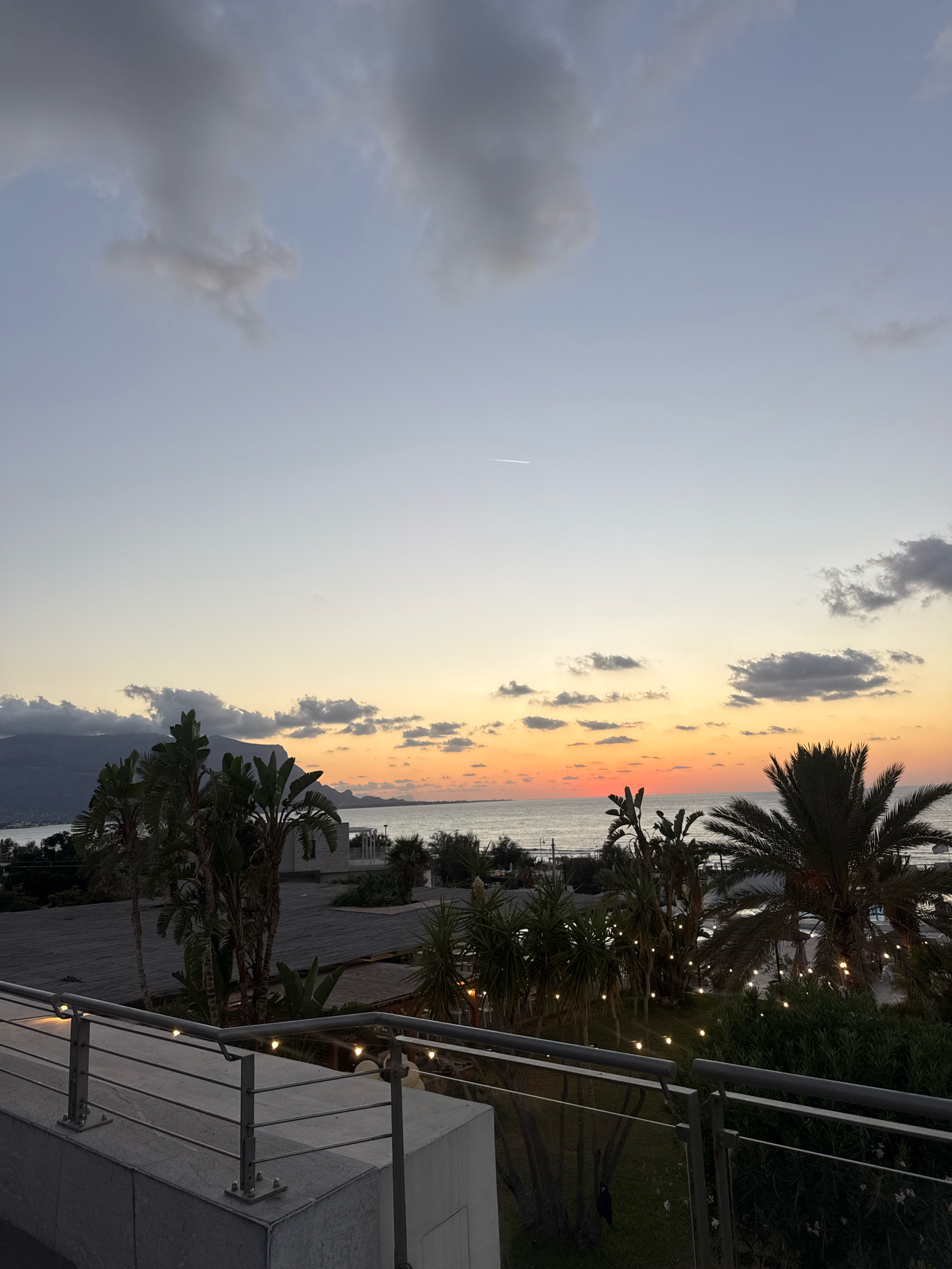 The image captures a sunset scene at a coastal location, likely in Isola delle Femmine, Italy. The sky transitions from a pale blue at the top to soft oranges and pinks near the horizon, indicating the late hour of the day. Darker, scattered clouds are visible, with a thin white contrail from an airplane stretching across the upper sky.
In the foreground, a modern-looking balcony or terrace features a metal railing with lights strung along it, casting a warm glow. Below the balcony, lush green foliage, including palm trees and other tropical plants, is illuminated by more string lights. A body of water, presumably the sea, stretches across the mid-ground, meeting the distant shoreline.
Further in the distance, a mountainous landscape is silhouetted against the colorful sky, with hints of buildings and structures visible along the coast. The overall atmosphere is serene and picturesque, characteristic of a Mediterranean evening. No people are visible in the frame. The scene is static, focusing on the natural beauty of the sunset and the surrounding vegetation.