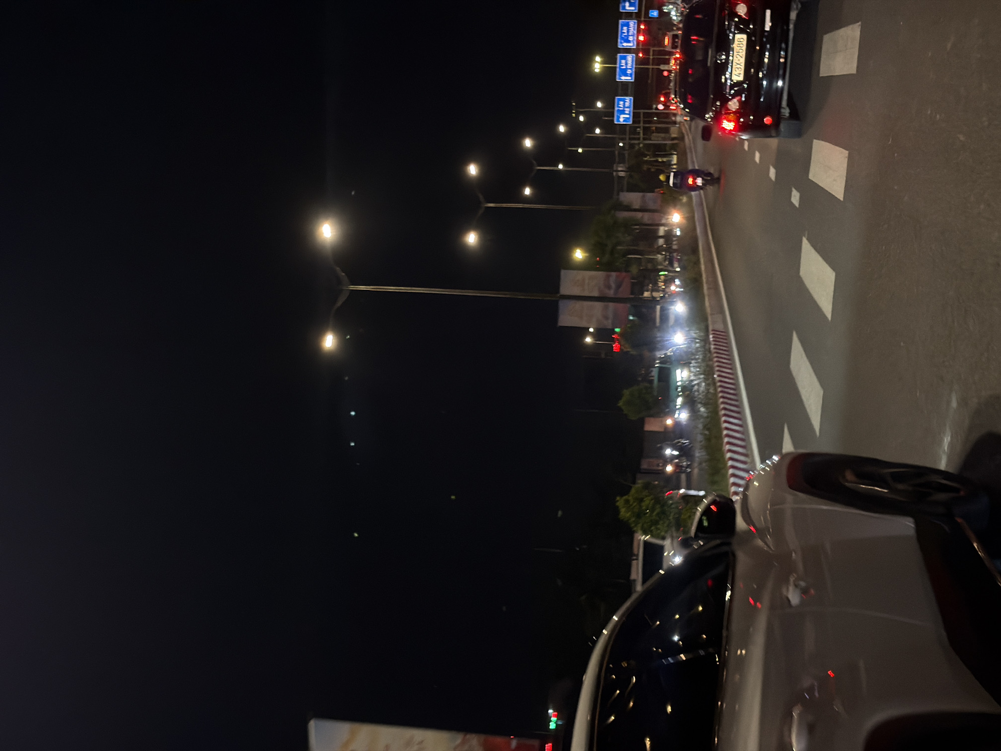 This is a night-time, street-level shot taken in Đà Nẵng, Vietnam, likely from a vehicle's perspective. The scene is dominated by the dark night sky, illuminated by streetlights and the headlights and taillights of passing vehicles. The road, marked with white lines and squares, suggests a multi-lane thoroughfare.
Several cars and a motorcycle are visible. In the foreground, the rear of a white car is prominent, its taillights reflecting the streetlights. Further down the road, a black car with license plate "43X-2586" is visible, its red taillights shining. A motorcycle with a rider wearing a yellow helmet is also present, navigating the traffic.
Road signs, in blue with white text, are mounted on poles, indicating traffic direction or lane information. Streetlights with multiple bulbs are spaced along the road, casting pools of light. Beyond the road, glimpses of buildings and illuminated signs can be seen, hinting at a commercial or urban area. The overall atmosphere is one of a busy city at night, with traffic flowing. The image is slightly tilted, giving a dynamic feel to the scene. The lighting conditions suggest it is well after sunset, with artificial lights being the primary source of illumination.