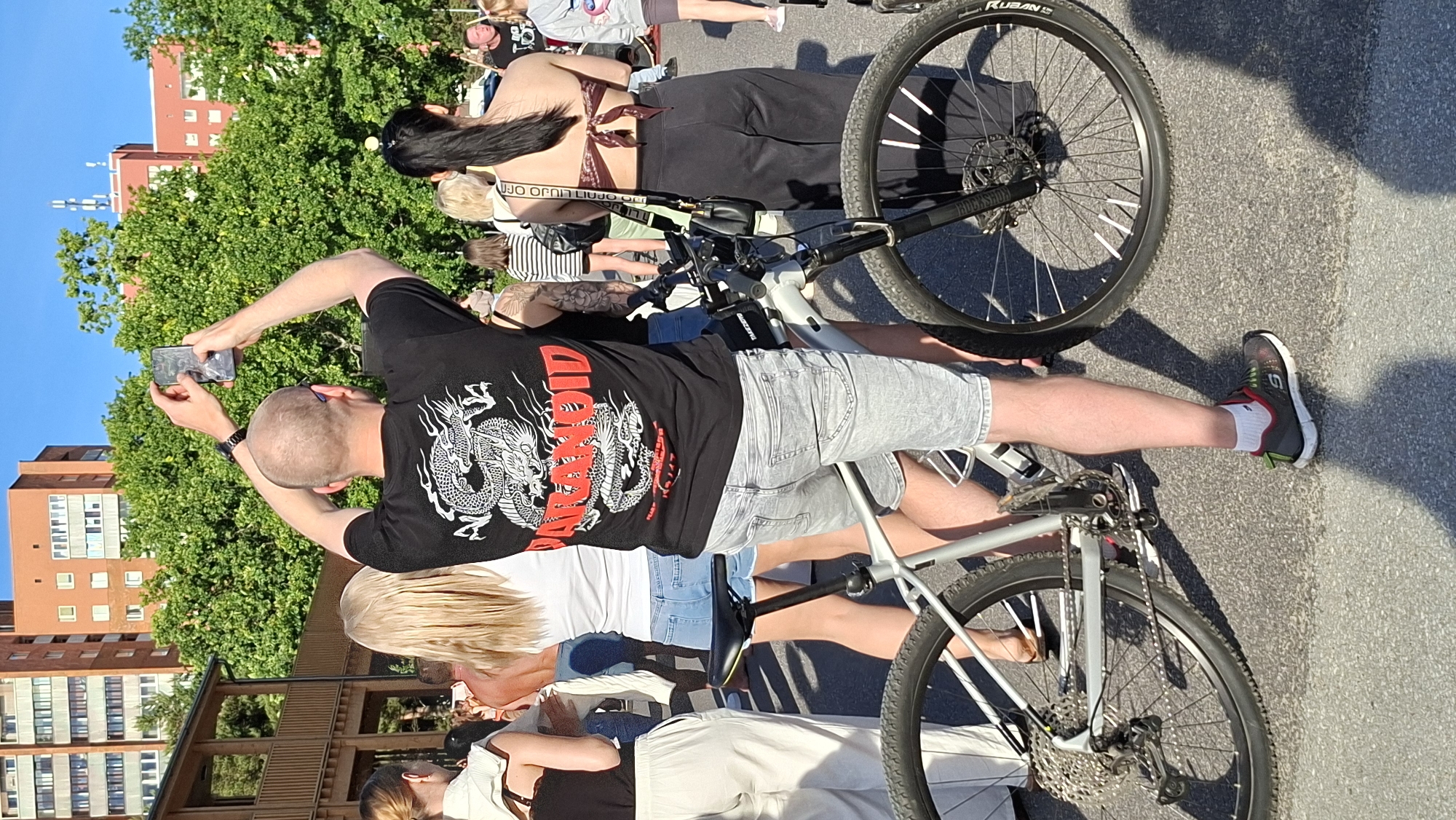 The image shows a group of people outdoors on a sunny day. The primary subject is a man in the foreground wearing a black t-shirt with a dragon graphic and light grey shorts. He is holding a smartphone in his left hand, seemingly taking a picture or video, with his arm outstretched. He is positioned next to a light-colored bicycle, with its front wheel visible. The bicycle has a black frame and a multi-gear system on the rear wheel.
Behind the man, several other people are visible. To his left, a woman with long dark hair is wearing a patterned bikini top and dark clothing. Further back, another woman with blonde hair is wearing a white top and jeans. The background features lush green trees and a tall, reddish-brown building with multiple windows. The scene appears to be a public space, possibly a park or a street, given the presence of the building and the asphalt ground.
The sunlight casts distinct shadows, indicating it is daytime. The bright lighting suggests fair weather. The visible text on the bicycle tire reads "RUBAN S". The overall mood seems casual and relaxed, with people gathered outdoors. The context provided places this scene in Tallinn, Estonia.