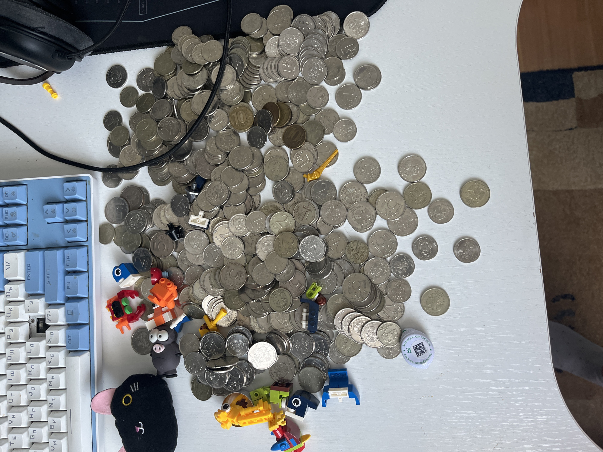 A flat lay shot shows a white desk surface cluttered with Russian coins, LEGO-like building toys, and a black plush cat toy with pink ears. A white and blue keyboard is visible in the upper portion of the image, with a black computer mouse and headphones on the right. The coins are scattered across the surface, some stacked, others spread out individually. The building toys include several small figures and pieces in bright colors like red, orange, yellow, and blue. The plush cat toy is black with a pink nose and ears, and two yellow eyes. The lighting is bright, casting minimal shadows. The scene does not contain any elements that can be used to identify a specific location within the city of Saratov, Russia.