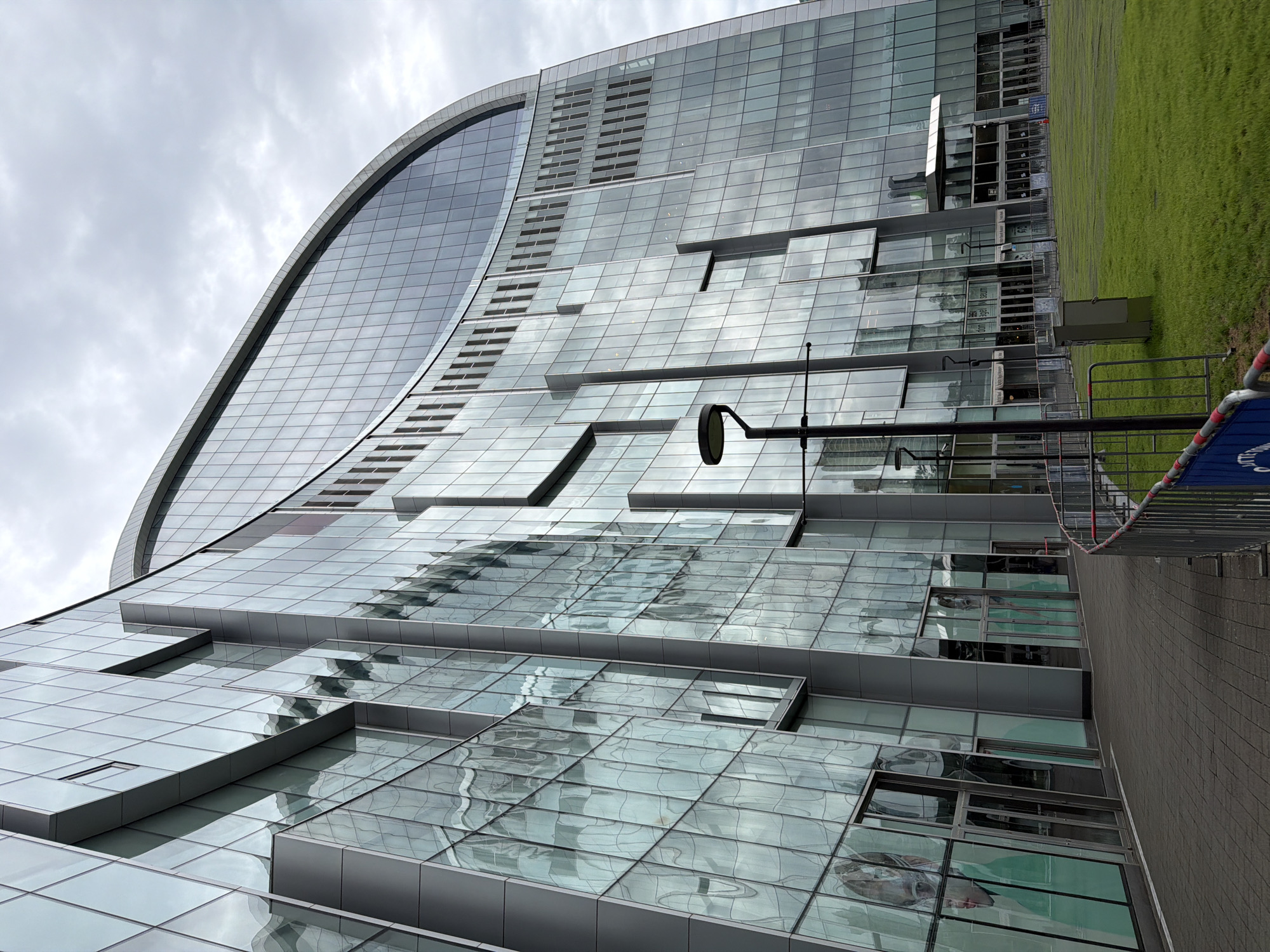 A wide-angle, eye-level shot captures the exterior of a modern, curved glass building, likely part of the Dongdaemun Design Plaza (DDP) in Seoul, South Korea, under an overcast sky. The building's facade is composed of numerous glass panels that reflect the sky and surrounding environment, creating a distorted, rippled effect. Geometric silver-toned frames delineate the glass sections. Several doors, some transparent with visible interiors, are set into the lower levels of the building. A paved walkway with a patterned surface leads along the building's base. To the right of the walkway, a manicured green lawn is bordered by a temporary metal fence. A black lamppost with a circular head stands prominently in the foreground, to the left of the building's entrance. The overall impression is one of contemporary architecture and urban design.