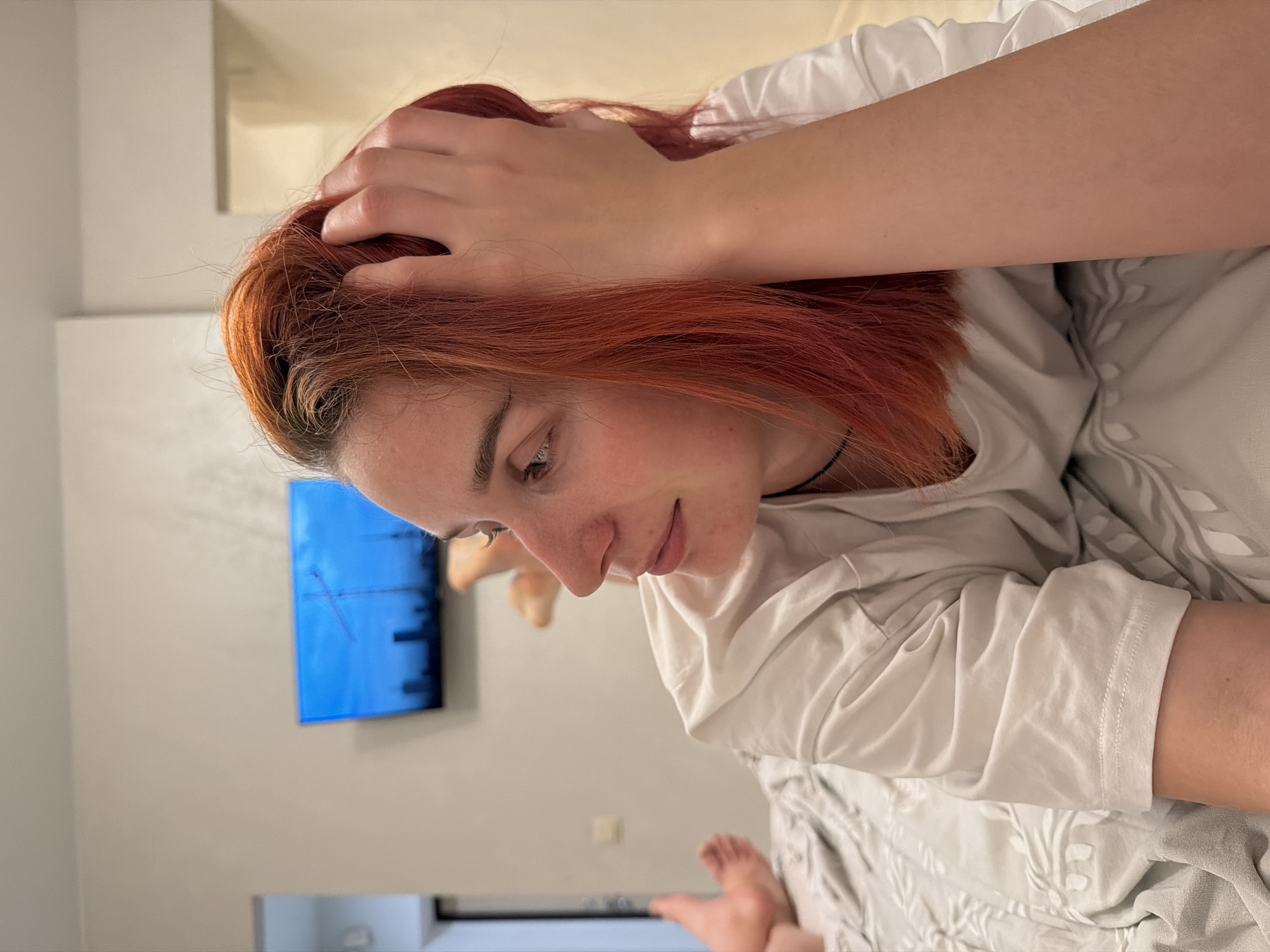The image depicts a woman with red hair lying on a bed. She is wearing a white t-shirt and her right hand is resting on her head. The background includes a wall-mounted television displaying a blue screen, a thermostat on the wall, and the end of the bed. Based on provided contextual information, the location is presumed to be in Naberezhnye Chelny, Russia.
