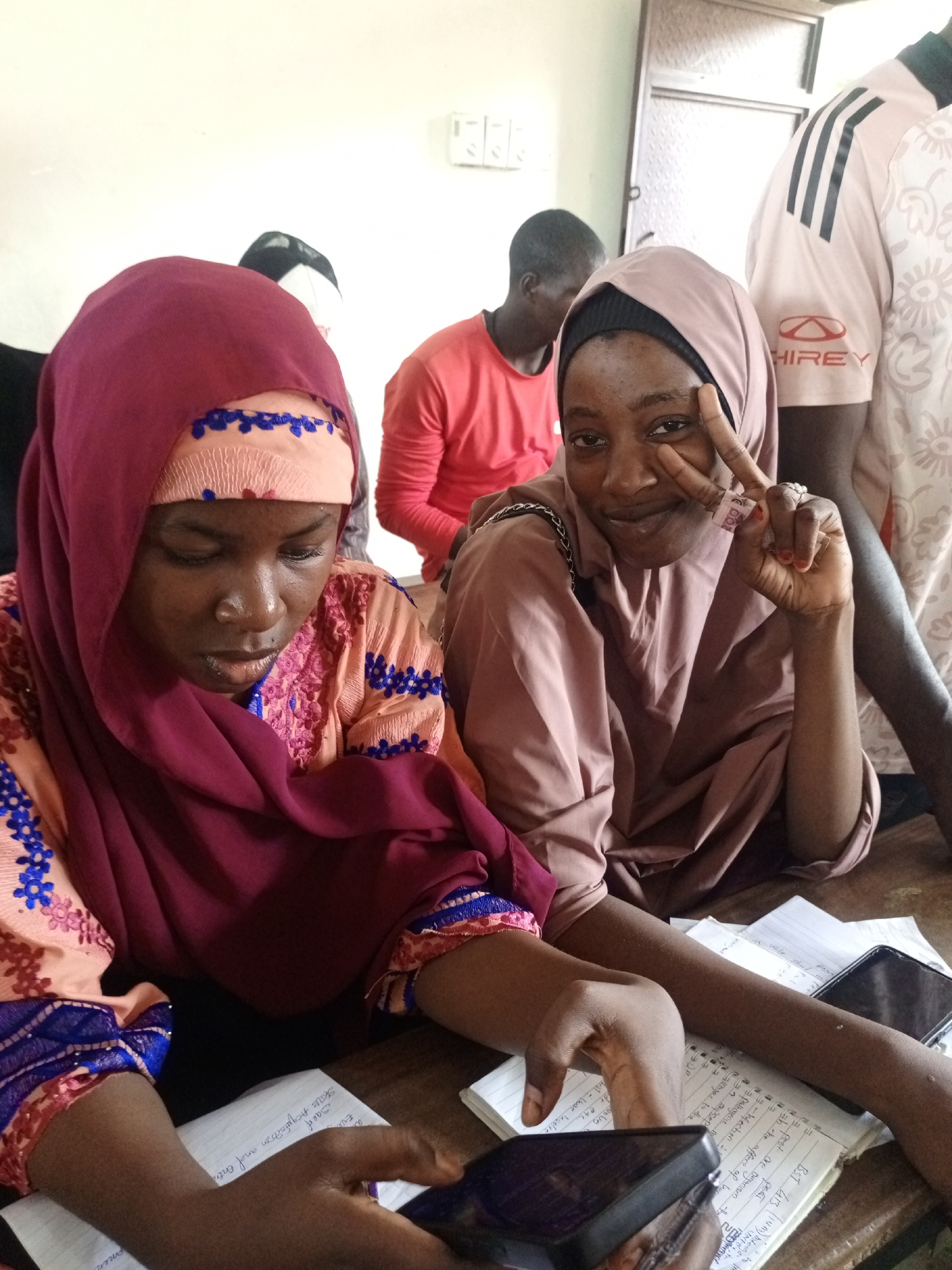 This image captures two young women seated at a table in an indoor setting, likely a classroom or study area in Keffi, Nigeria. The woman on the left, wearing a maroon hijab and a peach top adorned with blue embroidery, is focused downwards, appearing to interact with a mobile phone in her hands. Beside her, the woman on the right, dressed in a light brown hijab, smiles warmly and makes a peace sign with her right hand, which also holds a folded 200 Naira banknote.
In the background, two men are partially visible; one in a red t-shirt on the left, and another on the right wearing a light-colored t-shirt with black stripes and a "HIREY" logo. The scene features a plain white wall and what appears to be a door or cabinet.
On the table in front of the women are open notebooks containing handwritten notes. Notable visible text includes "BST 413," "PEST," "19-Infection," "pathogenice," "supsep," "Skaus Acquiston and Entre," and "pest are organian," suggesting academic study, possibly related to biology or pest management. Another mobile phone rests on the table. The lighting suggests it is daytime. The mood appears relaxed, with one subject posing cheerfully for the camera while the other is engaged in her activity.