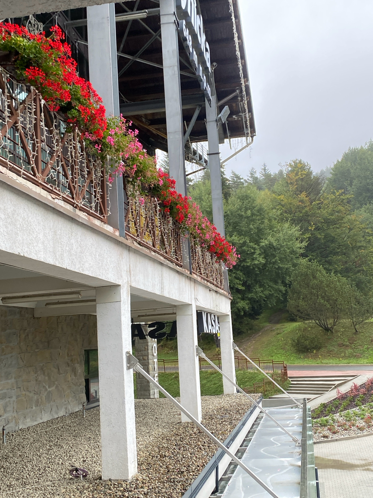 The image depicts the exterior of a building in Krynica-Zdrój, Poland, likely a ski resort or a similar mountain establishment, with the word "KASA" (which means "cash desk" or "ticket office" in Polish) visible on the building. The building features a white stucco facade with stone accents at the lower level. A balcony or terrace is adorned with abundant red and pink flowers in planters, along with decorative string lights. The background shows a lush green forest-covered hillside under a hazy, overcast sky. Stairs and pathways are visible leading up the hill. The foreground shows a gravel-covered area and a modern-looking transparent walkway with metal supports.