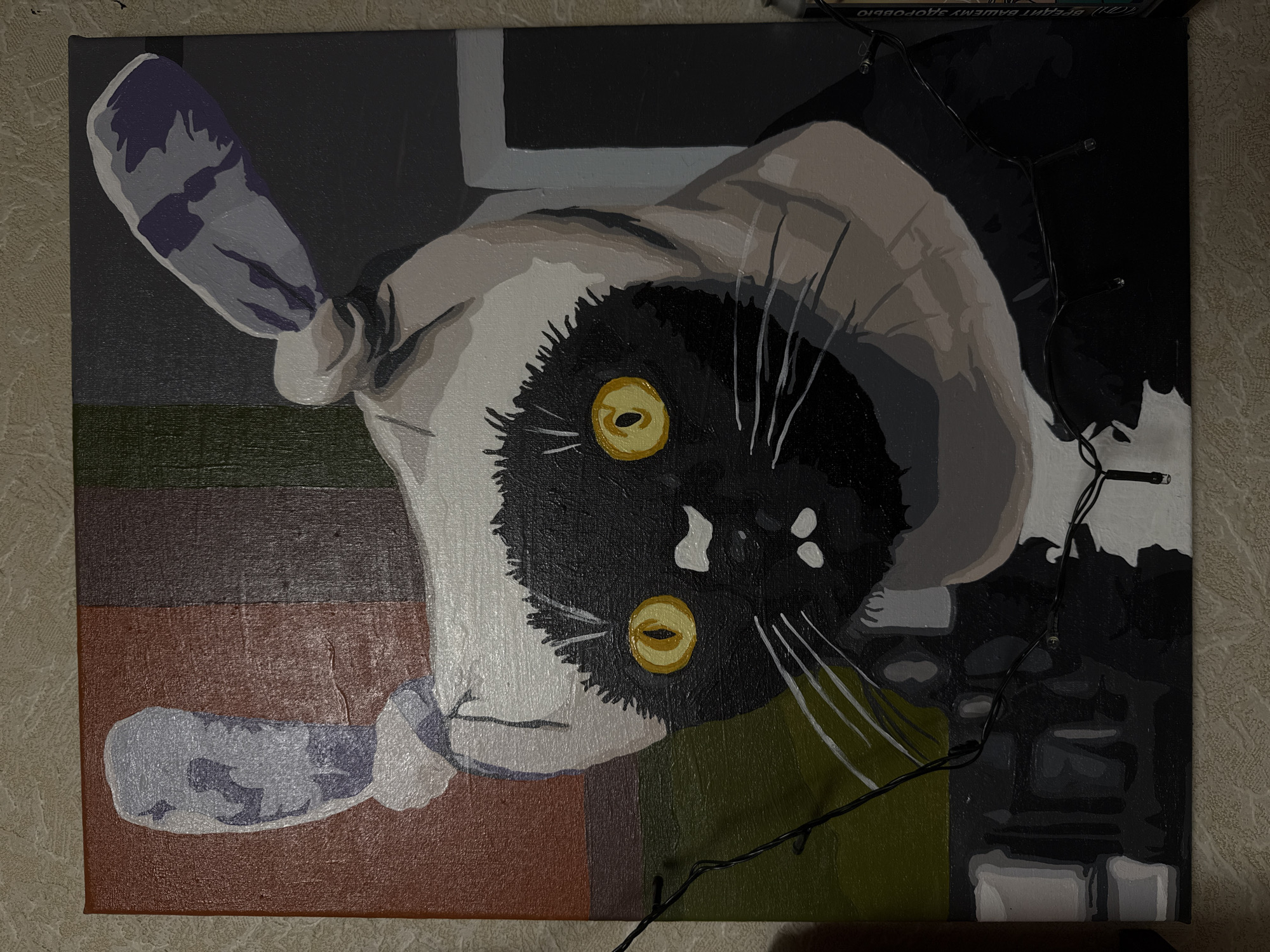 This is a painting of a black cat wearing a white and gray hat with two ear-like protrusions. The cat has large, yellow eyes and white whiskers. The background is abstract, with vertical bands of color including red, brown, green, and dark gray. There are also some horizontal shapes in shades of gray and white. In the foreground, a string of small, clear lights is draped across the lower part of the painting, with one light visible on the right edge of the canvas. The painting is on a textured surface, likely canvas, and appears to be unframed. The location is not discernible from the image.