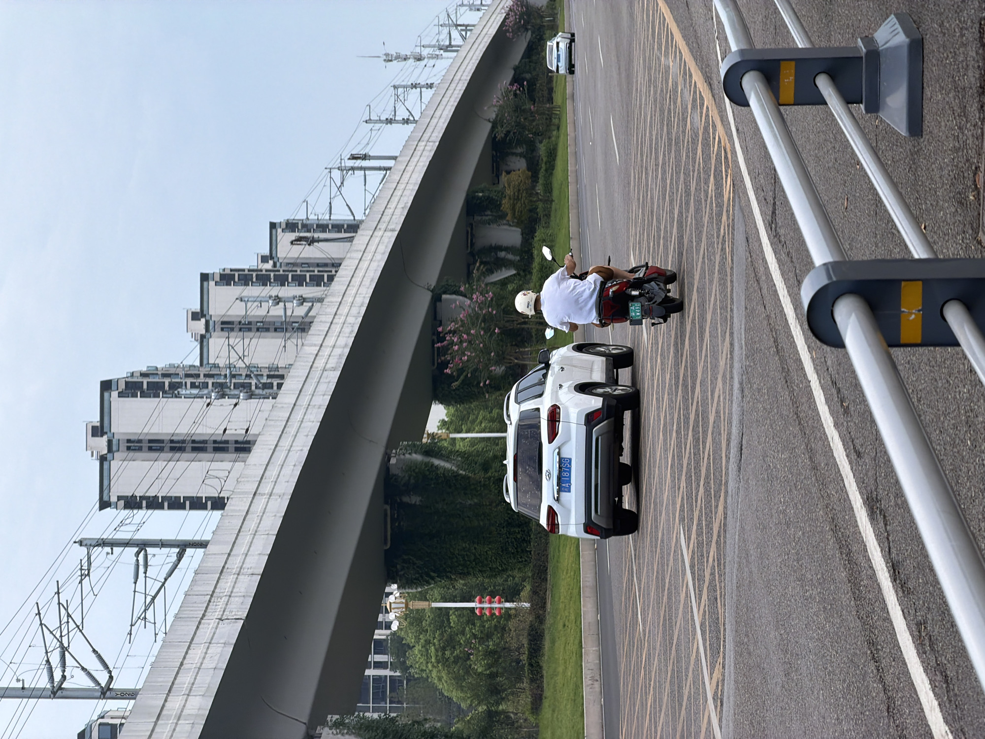 In Jiangjin, China, on a cloudy day, a white SUV is parked on the right side of a road. A person is riding a red electric scooter in the same lane, heading away from the viewer. In the background, a multi-level overpass with overhead train wires stretches across the scene. Several tall, modern buildings are visible behind the overpass. The road is marked with yellow and white lines, and a metal guardrail is in the foreground. Green trees and shrubbery line the side of the road.