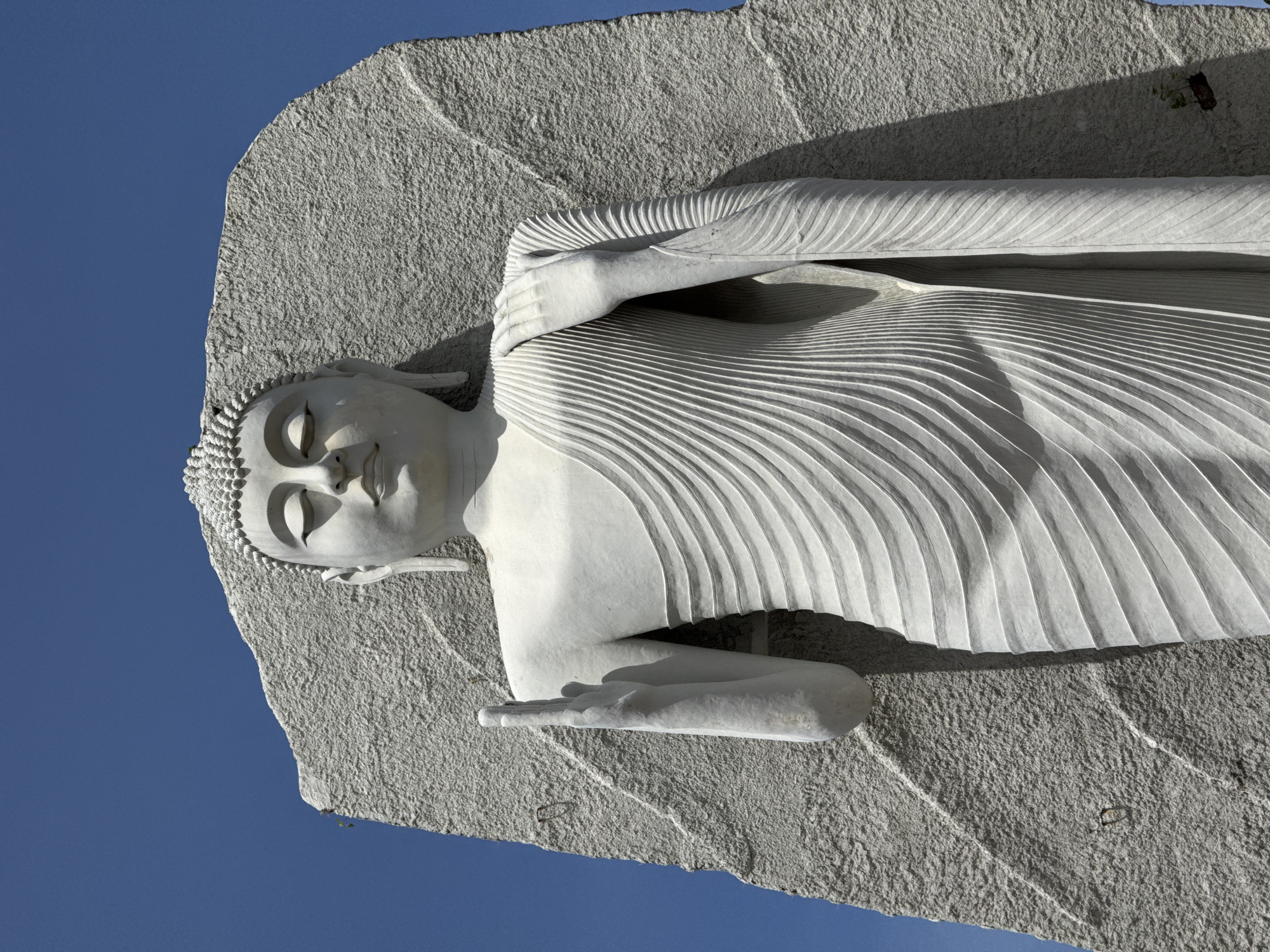 The image shows a close-up, low-angle shot of a large, white Buddha statue carved from stone, set against a clear blue sky. The statue is depicted standing, with its eyes closed and a serene expression on its face. Its right hand is raised with the palm facing outwards, in a gesture of blessing or protection, while its left hand rests on its shoulder, draped with intricate, flowing robes. The stone background behind the statue is rough and textured, contrasting with the smooth finish of the sculpture. The statue is located in Dondra, Sri Lanka.