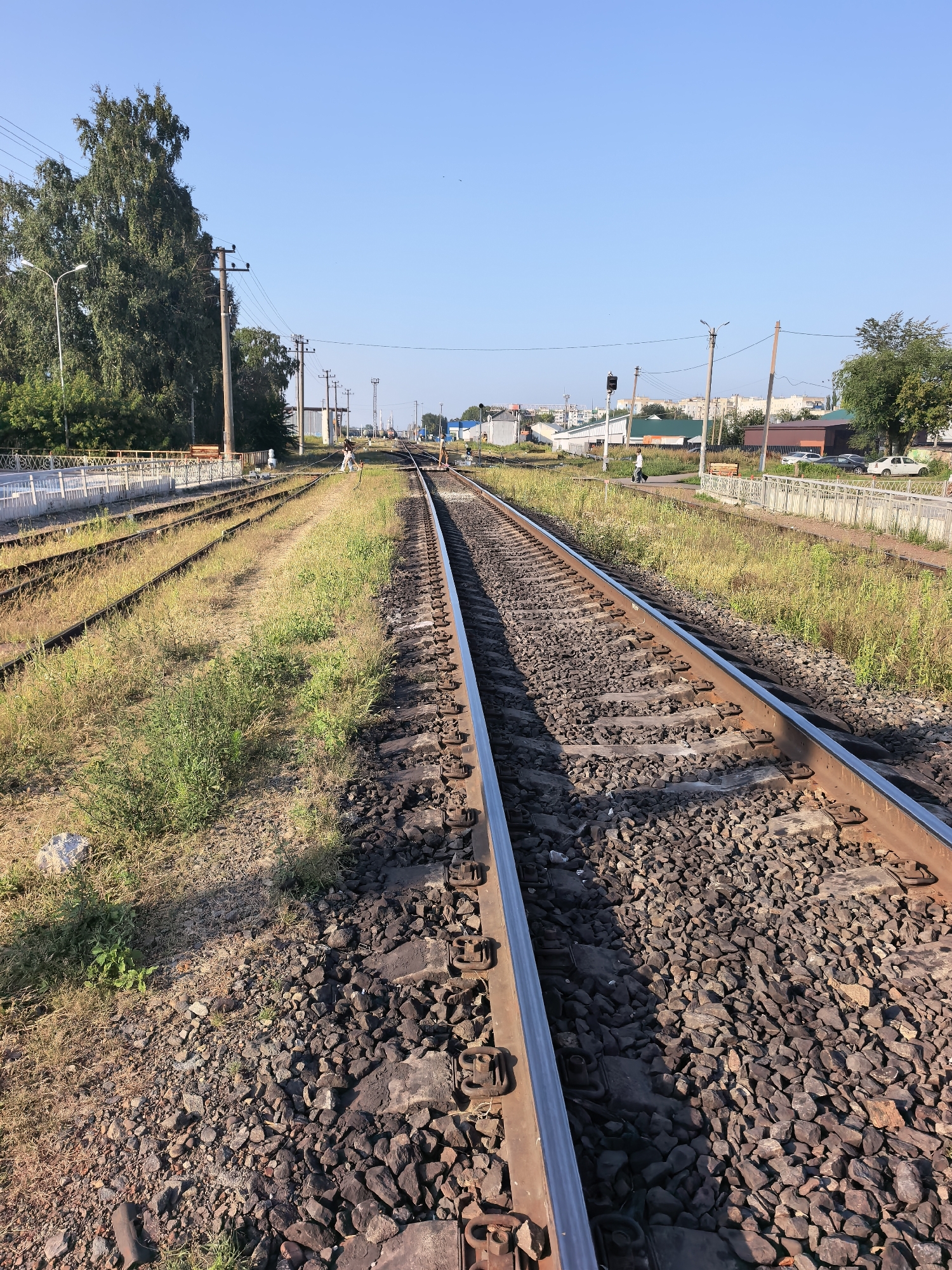The image shows a railway track in the city of Sterlitamak, Russia, on a clear, sunny day. The tracks run towards the horizon, with two parallel sets of tracks visible in the foreground. The area surrounding the tracks is covered in gravel and dry grass. In the background, there are buildings, trees, power lines, and a few cars parked. Two individuals are visible on or near the tracks, one in the middle ground walking away from the camera, and another further in the distance near the buildings. The scene appears to be an active railway station or yard, with the characteristic infrastructure of such a location. The lighting suggests it is either early morning or late afternoon.