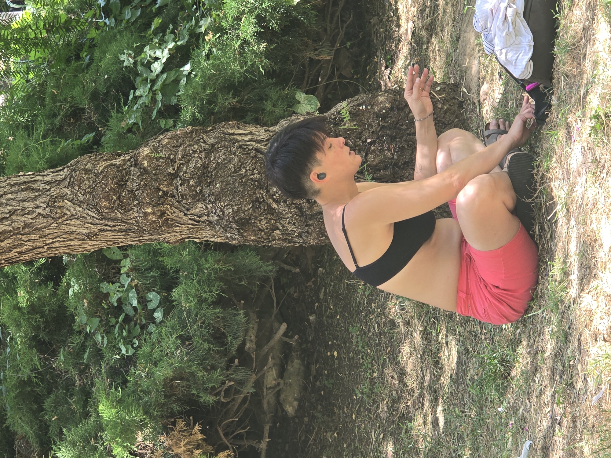 A person with short dark hair and earbuds in their ears is sitting on the ground next to a tree. They are wearing a black bra and pink shorts. The person is holding something in their right hand and looking down at it. There is a bag with clothes next to them. The ground is covered in grass and dirt. The background is filled with green bushes and branches. The scene appears to be in a park.