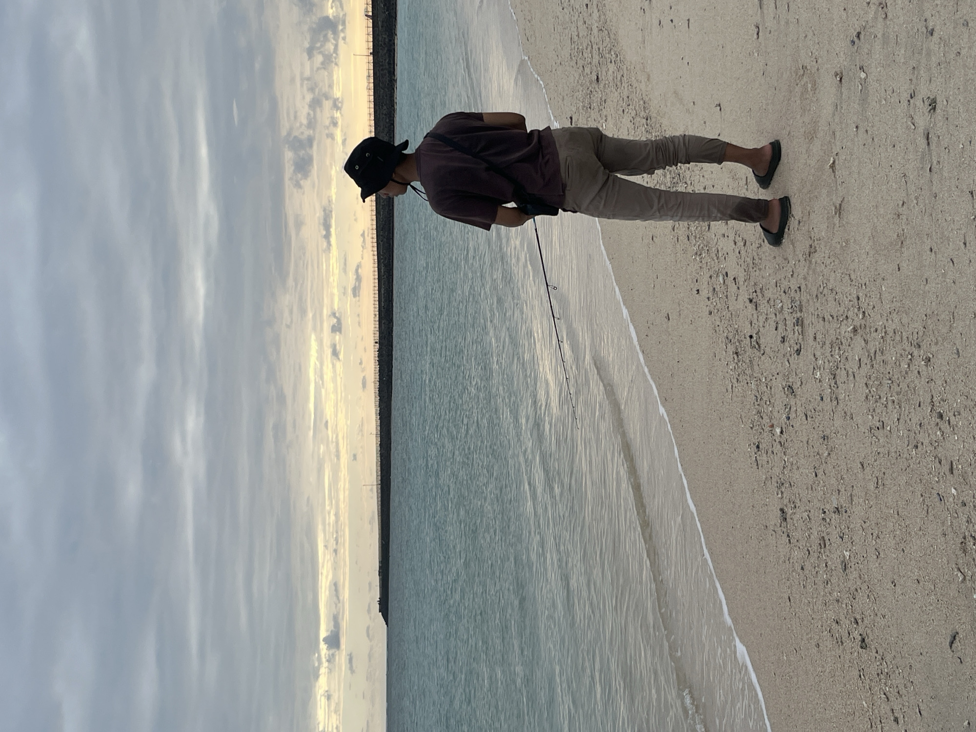 A lone figure stands on a sandy beach in Kuta, Indonesia, fishing in the shallow ocean waters. The person, dressed in a brown t-shirt, beige pants, and a black bucket hat, is facing away from the camera, holding a fishing rod. The ocean is a calm, turquoise color, with small waves lapping at the shore. The sky above is overcast with streaks of yellow and orange from the setting sun. In the distance, a breakwater extends into the sea, lined with a fence. The beach itself is composed of fine, light-colored sand, scattered with small shells and pebbles. The scene is serene, with the sounds of the waves and the gentle breeze creating a tranquil atmosphere.