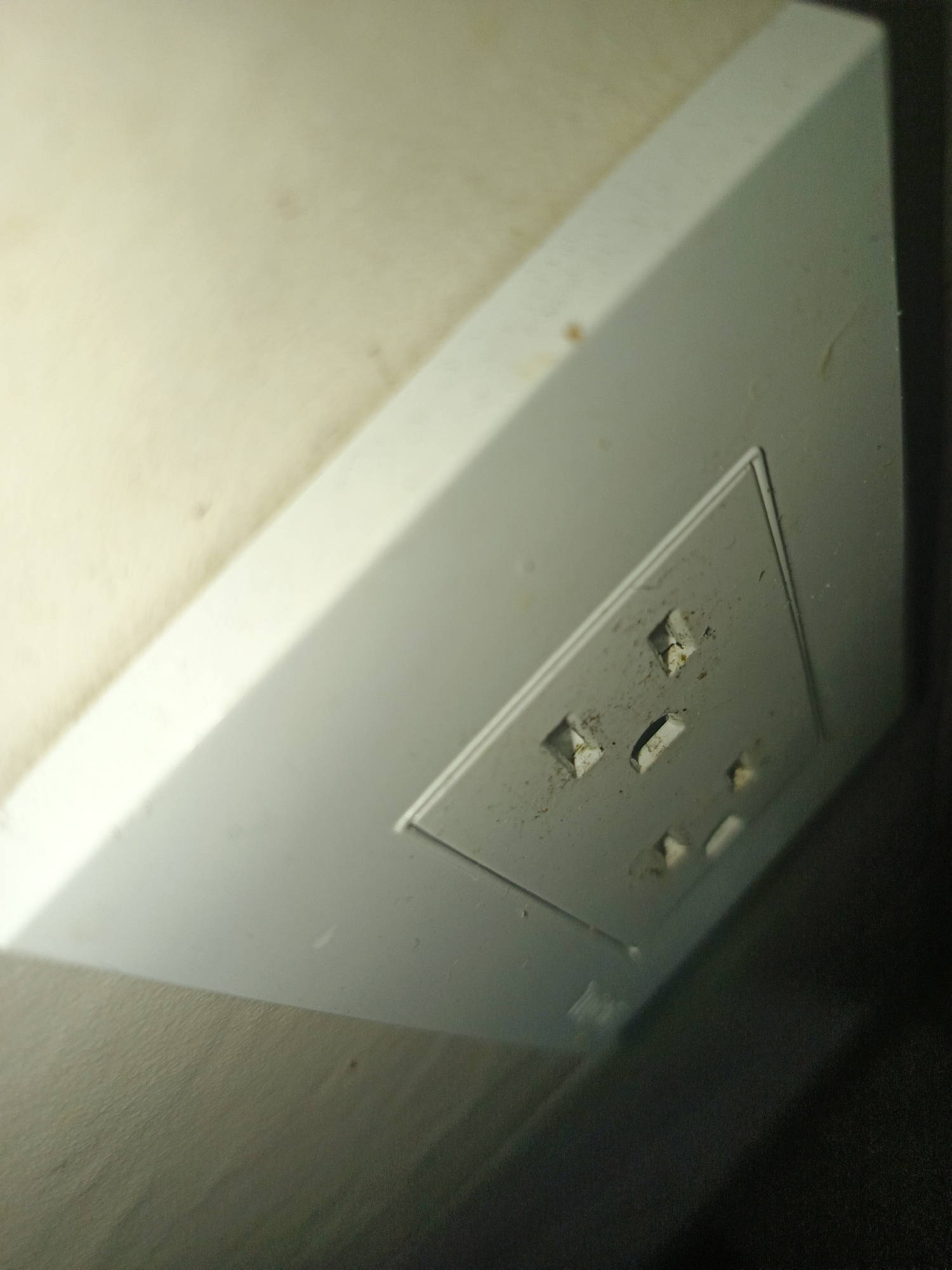 A close-up shot of a dirty white electrical outlet in what appears to be a wall. The outlet is recessed within a slightly larger white panel. The outlets show signs of dust and grime accumulation. The wall appears to be cream or off-white. To the right of the outlet is an area that is dark and out of focus, possibly indicating the edge of a wall or another surface. The overall lighting is dim, with the outlet appearing to be slightly angled toward the camera. The image is undated, but has indications of a very dirty electrical outlet in a residential setting. The location is Los Robles, Venezuela.