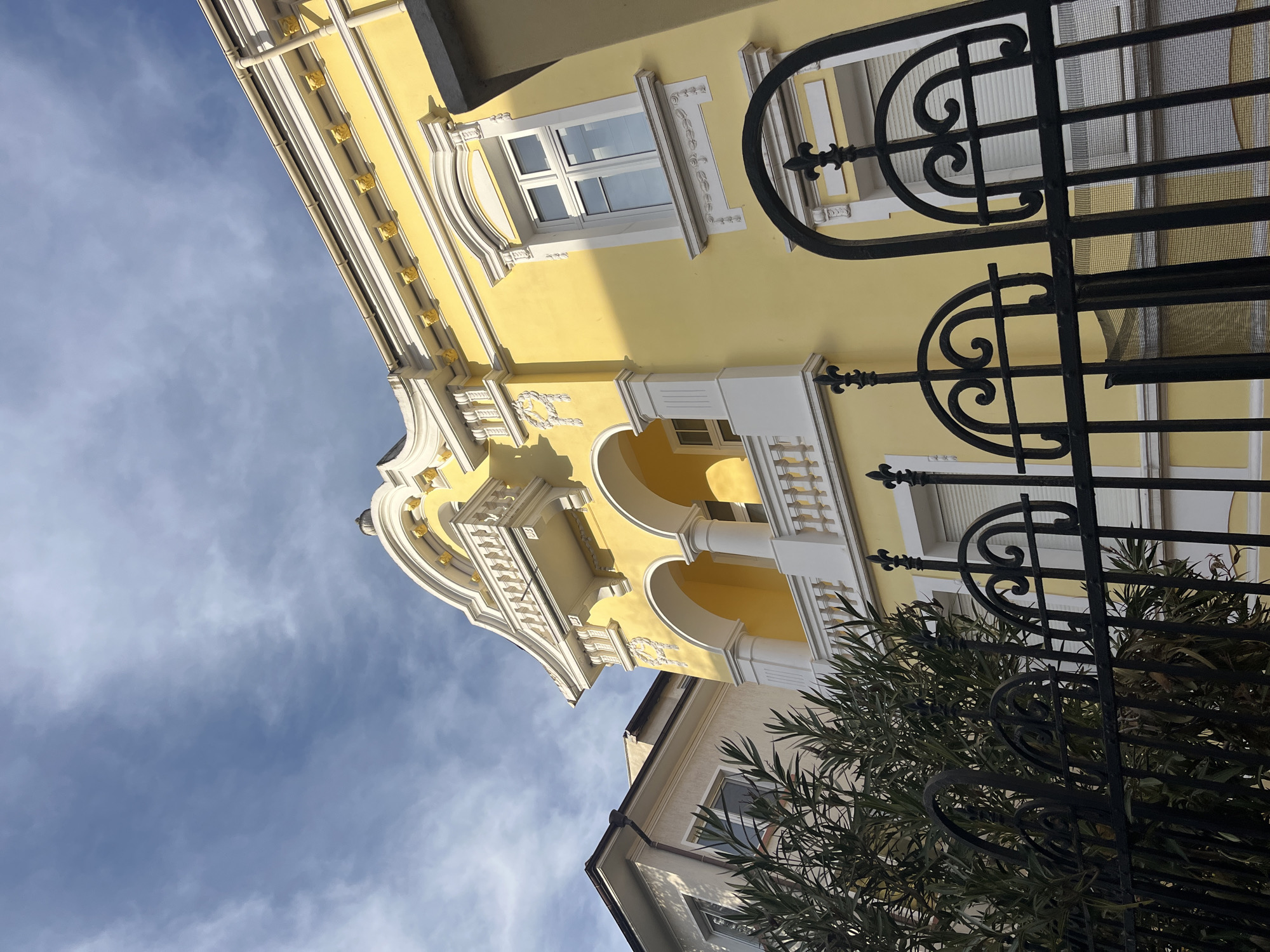 A low-angle shot captures a yellow building with ornate white architectural details, including arched balconies and decorative trim, set against a blue sky with scattered clouds. A black wrought-iron fence with decorative curves is prominently featured in the foreground. The branches of a tree obscure part of the fence and the building behind it. The building appears to be in Burgas, Bulgaria, based on architectural styles commonly found in the city.