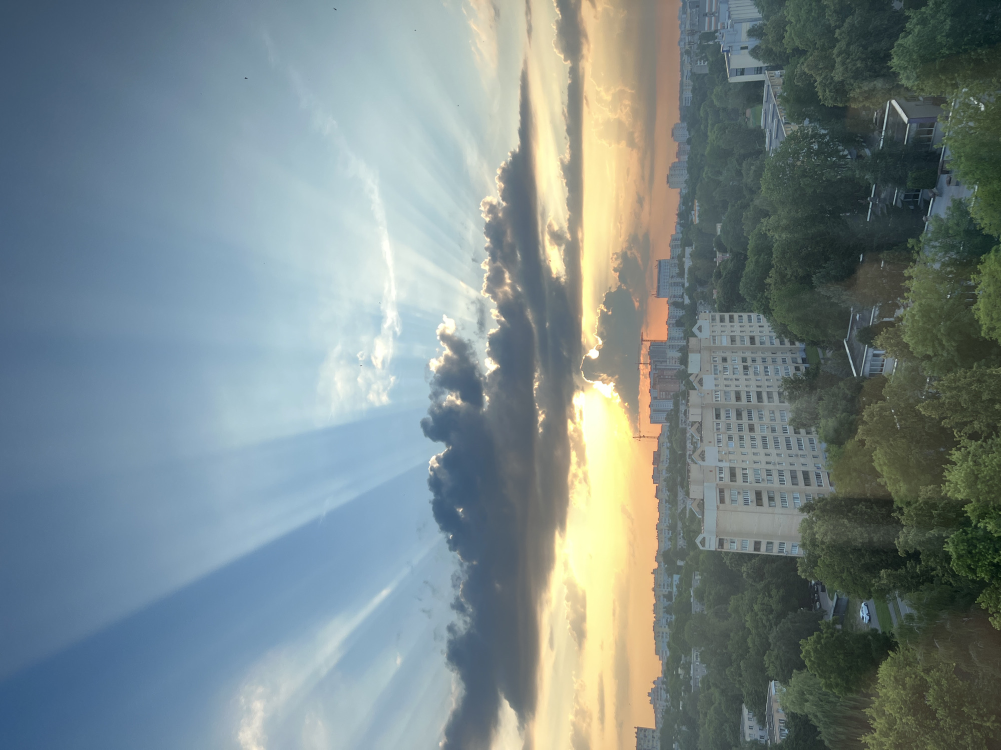This is a vertical, eye-level shot of a cityscape at sunset, captured from a high vantage point, likely a building. The scene is dominated by a dramatic sky with rays of sunlight piercing through clouds, creating a striking contrast between light and shadow. On the left, the sky is a clear blue, transitioning to a vibrant yellow and orange near the horizon on the right, indicating sunset. Dark, heavy clouds are positioned centrally, partially obscuring the sun, which is casting luminous rays.
Below the sky, a dense urban landscape unfolds. Numerous apartment buildings, characterized by their multi-story structure and numerous windows, are interspersed with lush green trees. The foliage suggests a summer or late spring season. The overall impression is one of a serene, yet dynamic evening in a city. The orientation of the image is unusual, as the cityscape appears to be at the top and the sky at the bottom, suggesting the photo was taken vertically or rotated. The image's composition evokes a sense of awe and tranquility, highlighting the beauty of a natural phenomenon over an urban setting. No visible text is present in the image.